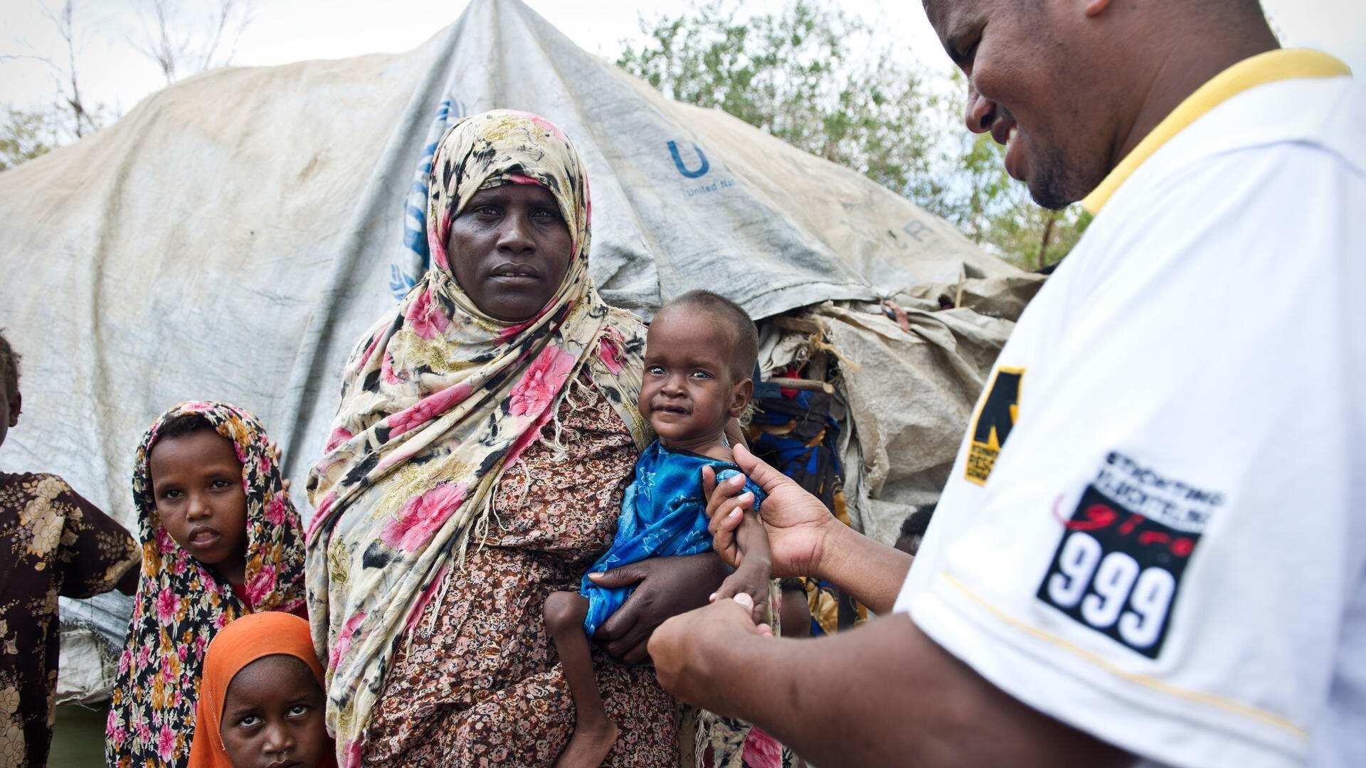 Eine Mutter hält ihr Baby im Arm. Ein Mitarbeiter von IRC untersucht das Baby. Im Hintergrund steht ein Zelt.