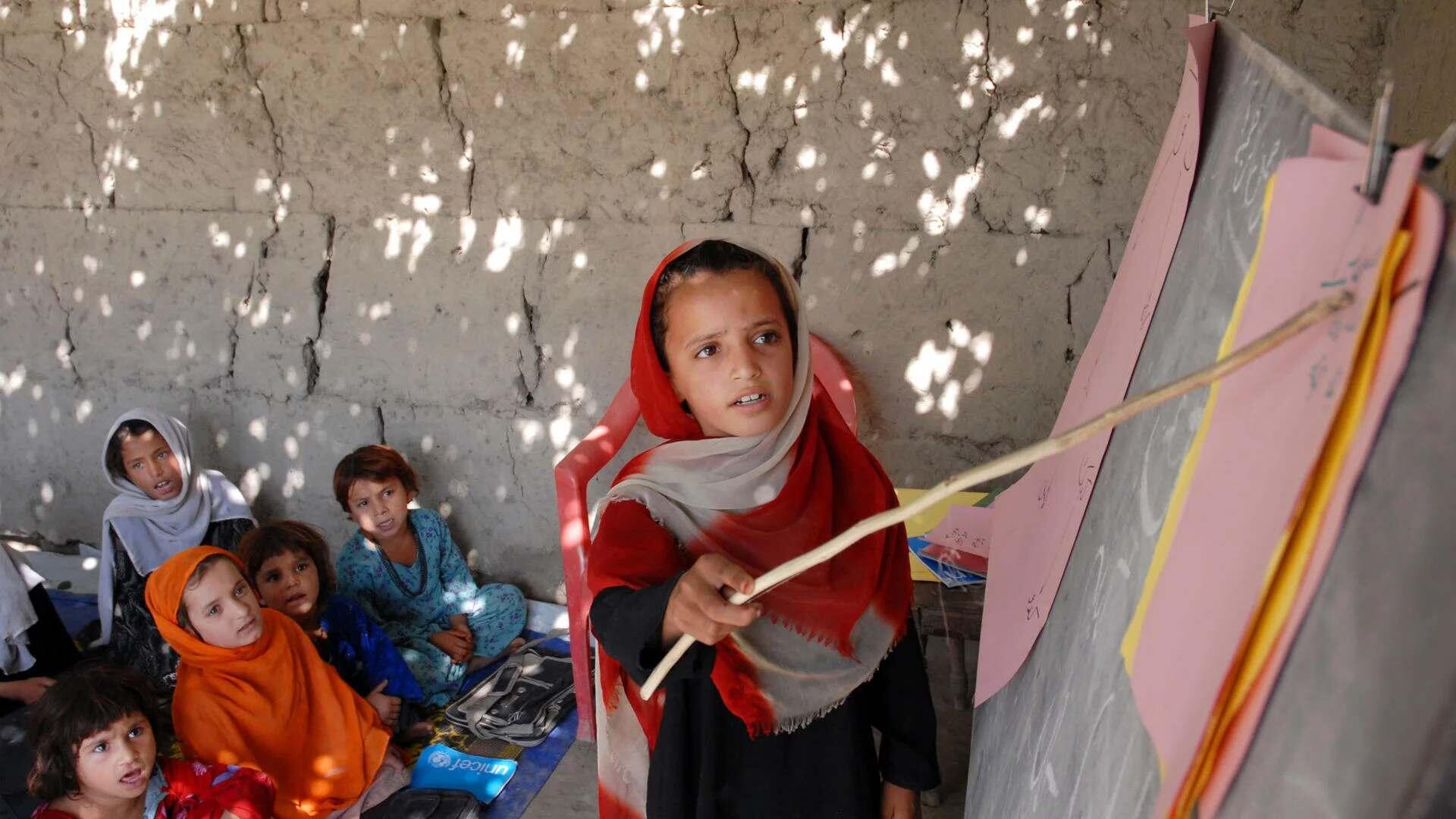 Ein Mädchen zeigt mit einem Stock auf eine Tafel, im Hintergrund sind ein paar Kinder, die ihr zuschauen.