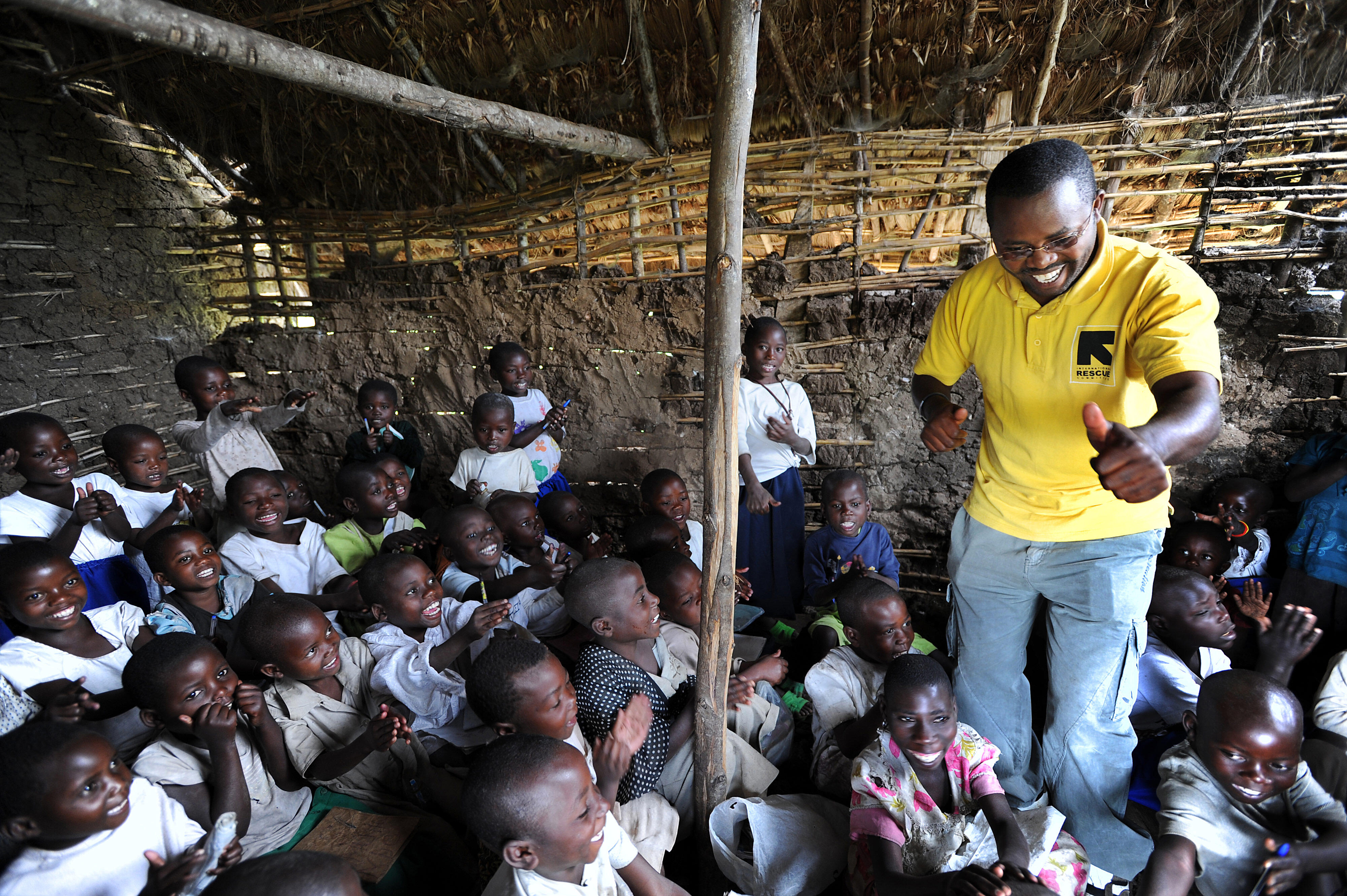 A smiling aid worker stands amidst a classroom of children, leading them in a song