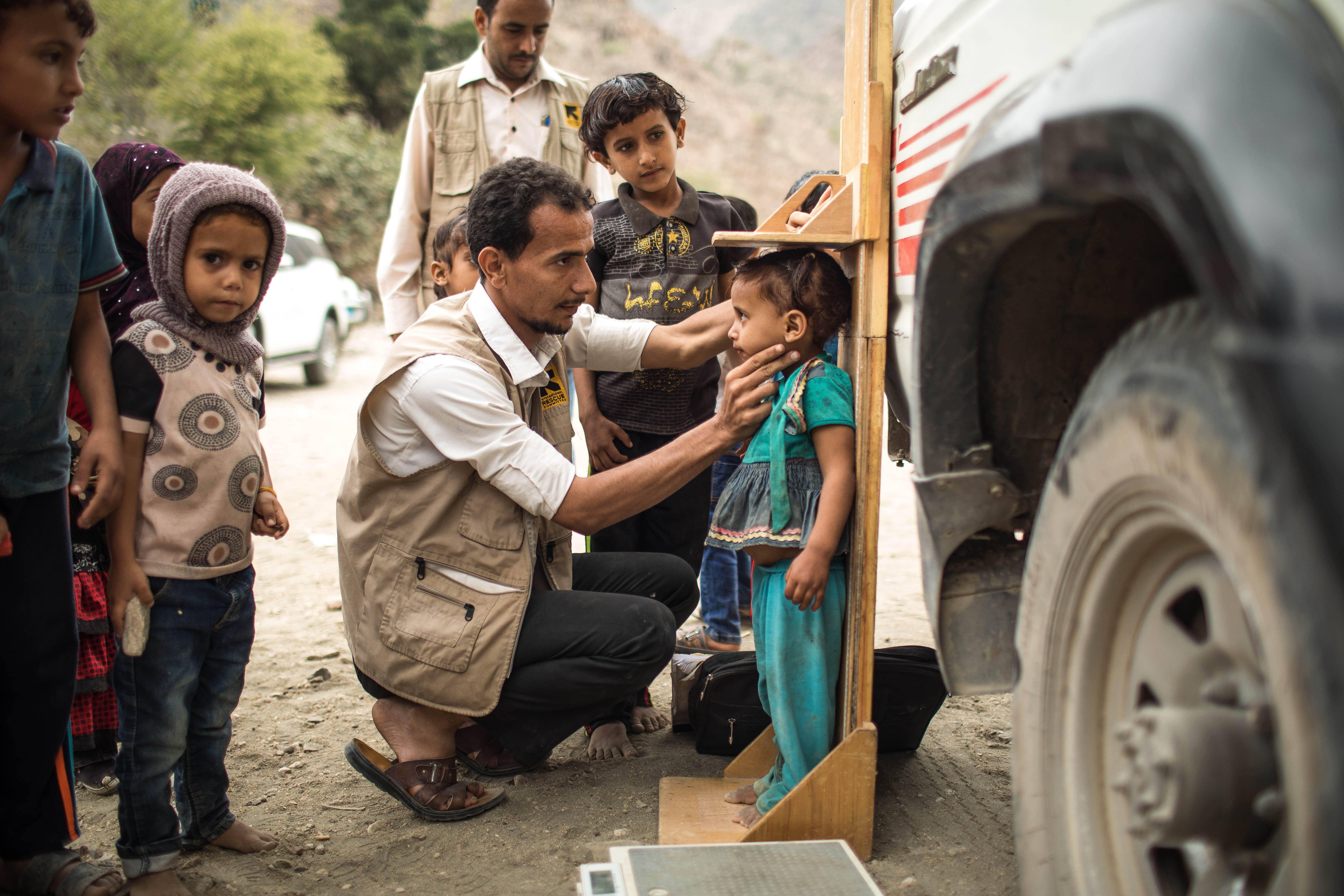 IRC 이동진료소의한남성의료진이예멘의외딴산지마을에서영양실조 징후를보이는아동을진찰하고, 다른아동들이그장면을지켜보고있습니다.