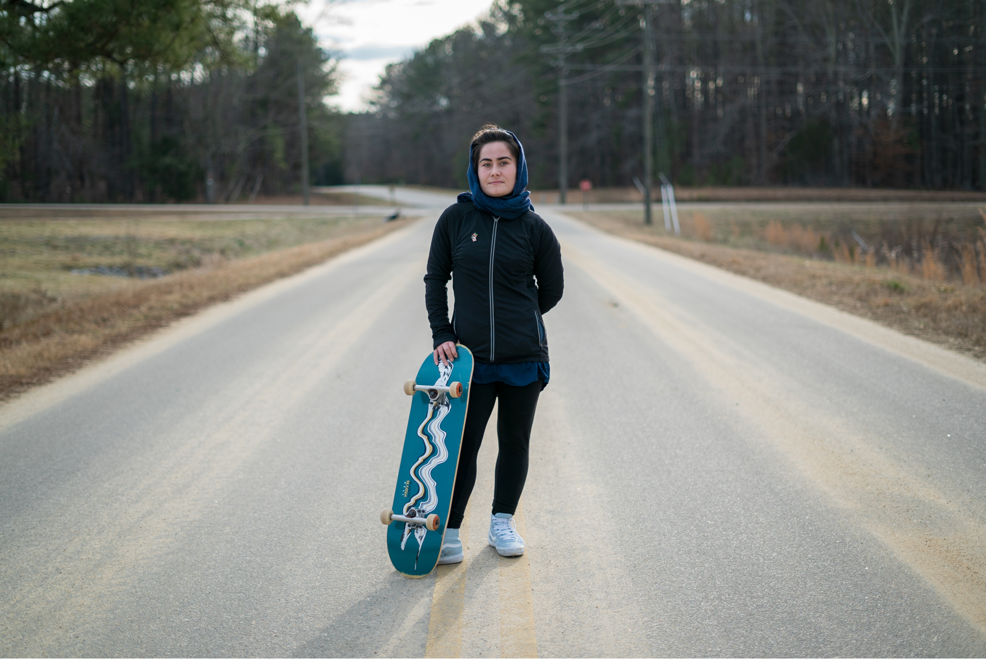 A 22 year old woman stands in the middle of a road holding a skateboard