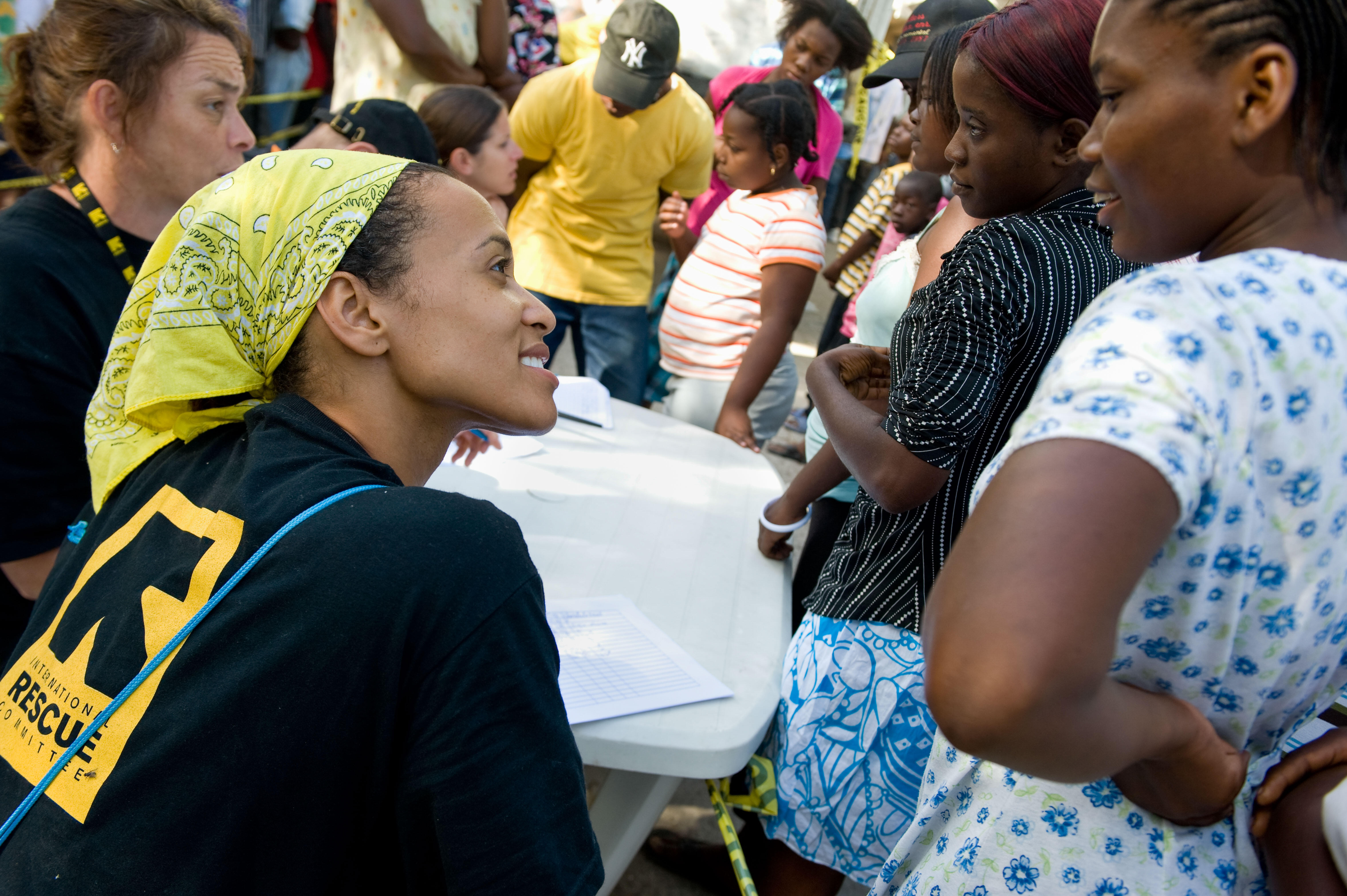 A woman wearing an IRC shirts smiles while interacting with a group of people waiting for humanitarian support.