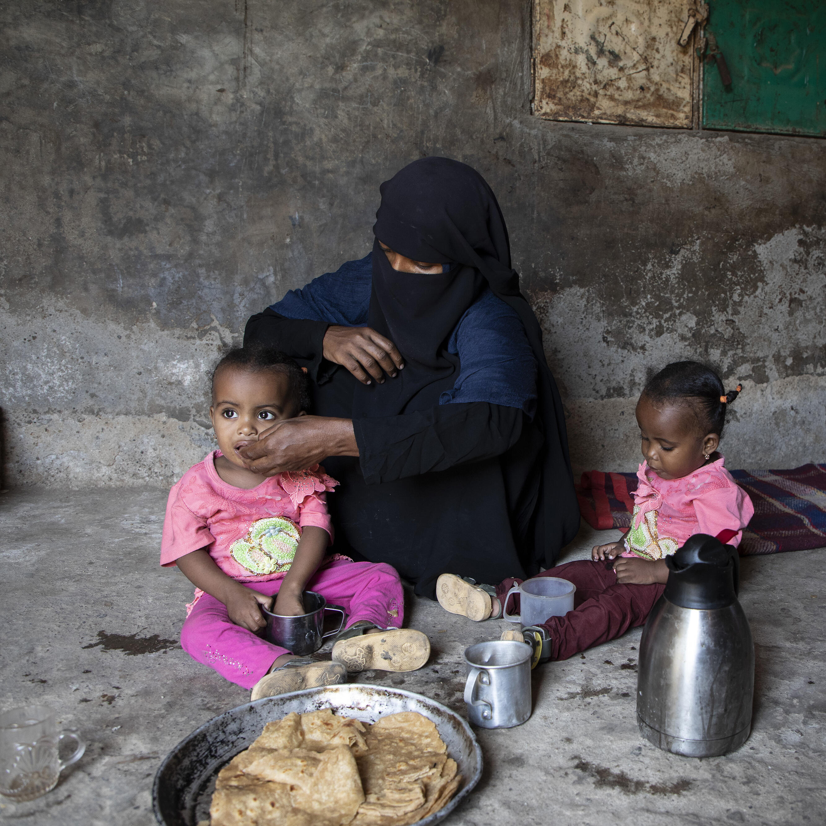 A mother siting  on the floor feeding her two children.