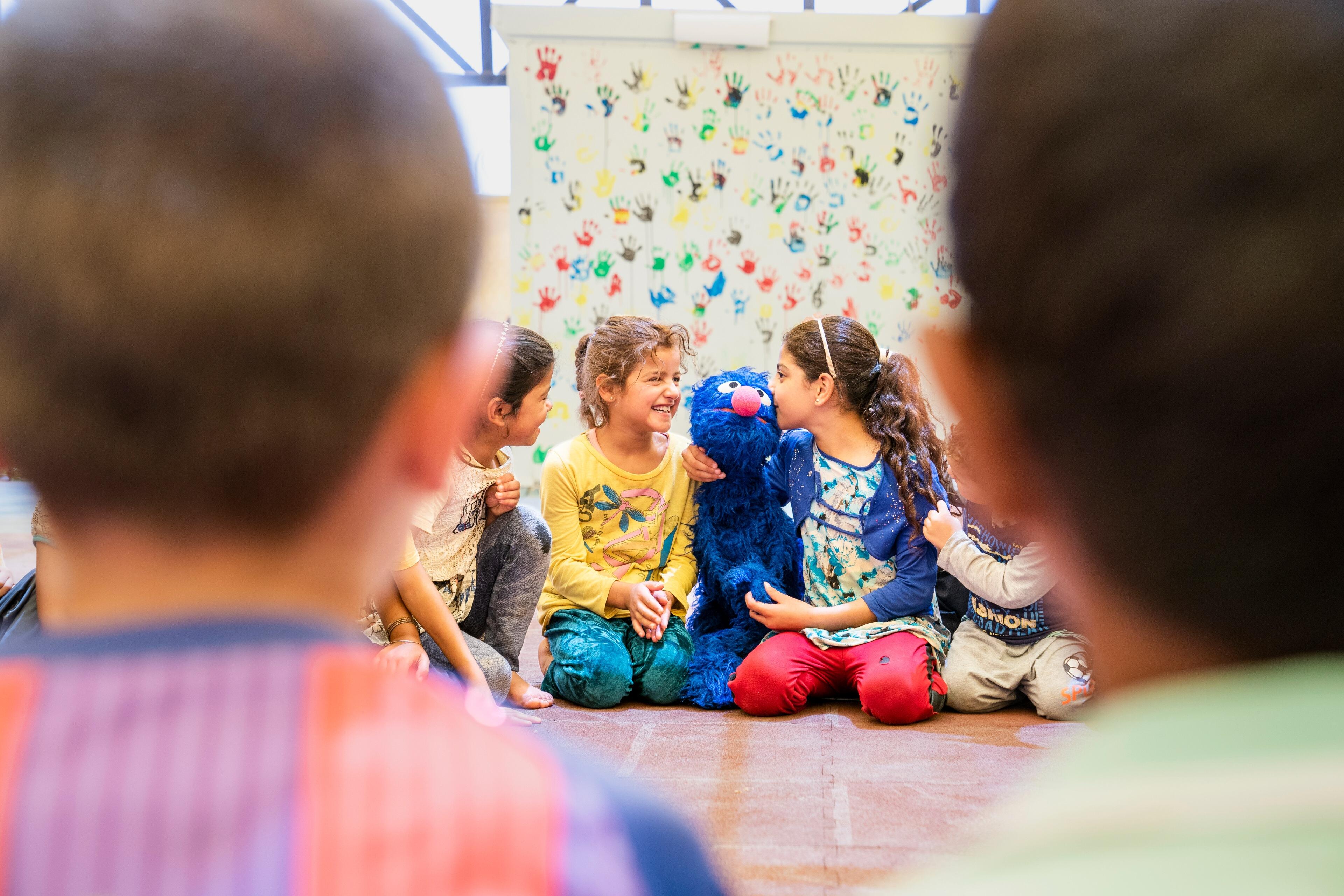 Refugee children sit on the ground in a circle playing witha puppet of the Sesame Street character Grover, a blue monster.