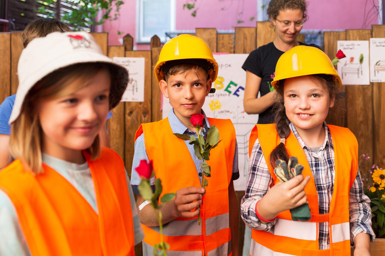 Ein Junge und zwei Mädchen halten Rosen in ihren Händen und präsentieren ihr Hochbeetprojekt. Alle drei tragen eine Warnweste und eine Kopfbedeckung.