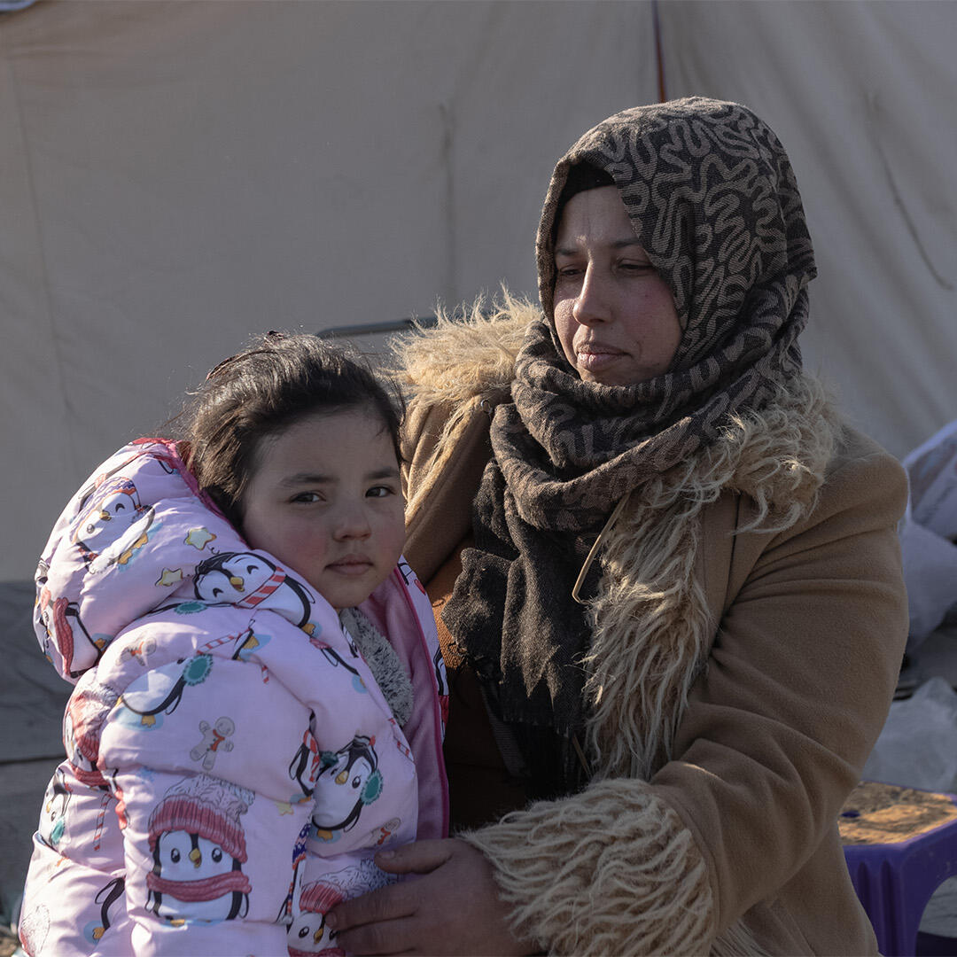 Latifa is holding her 4 year old, Jalian. They are sitting in front of a tent in a camp for displaced people in Türkiye.