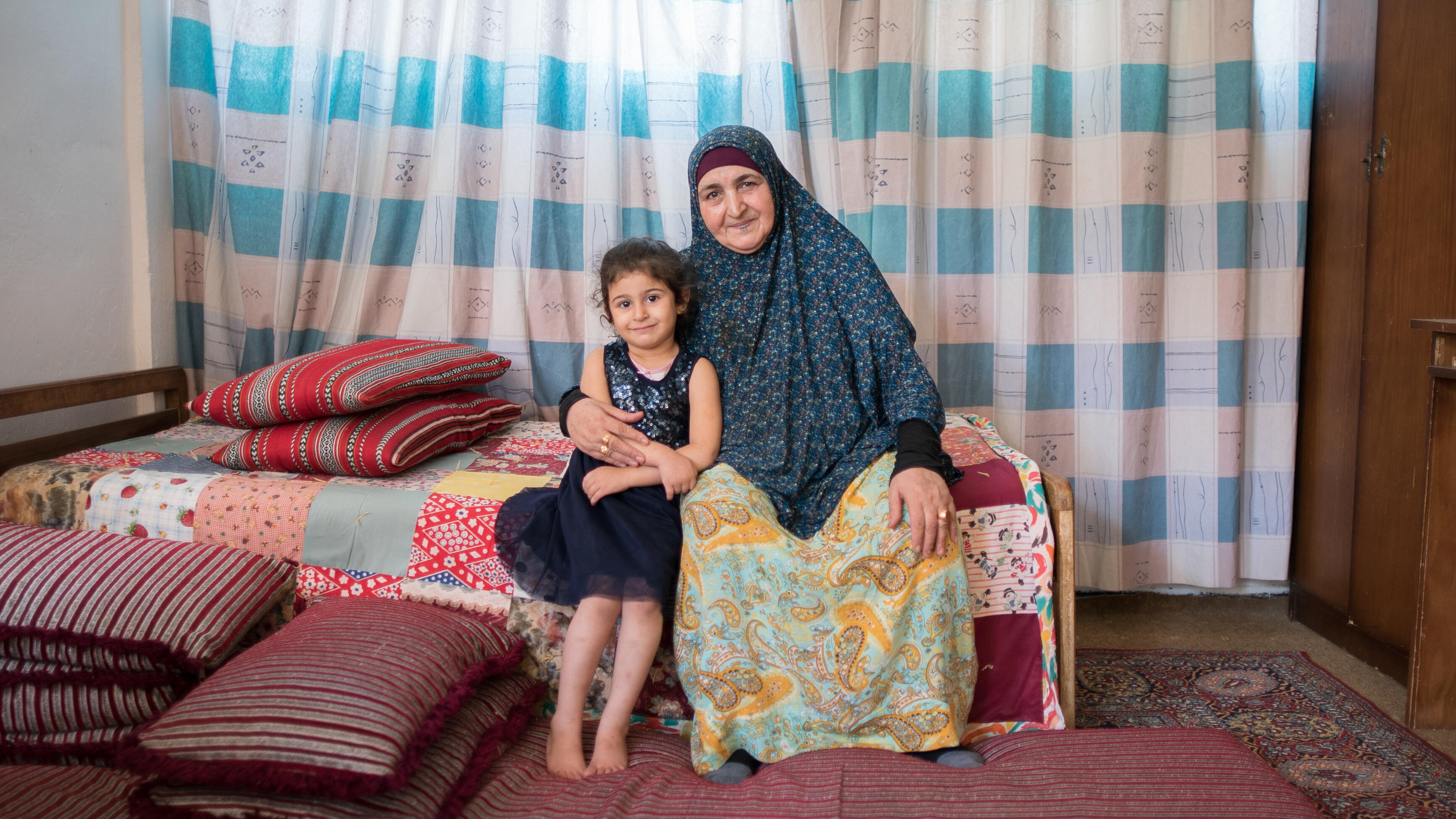 A young girl and her grandmother sit at the edge of a bed and pose for a photo.
