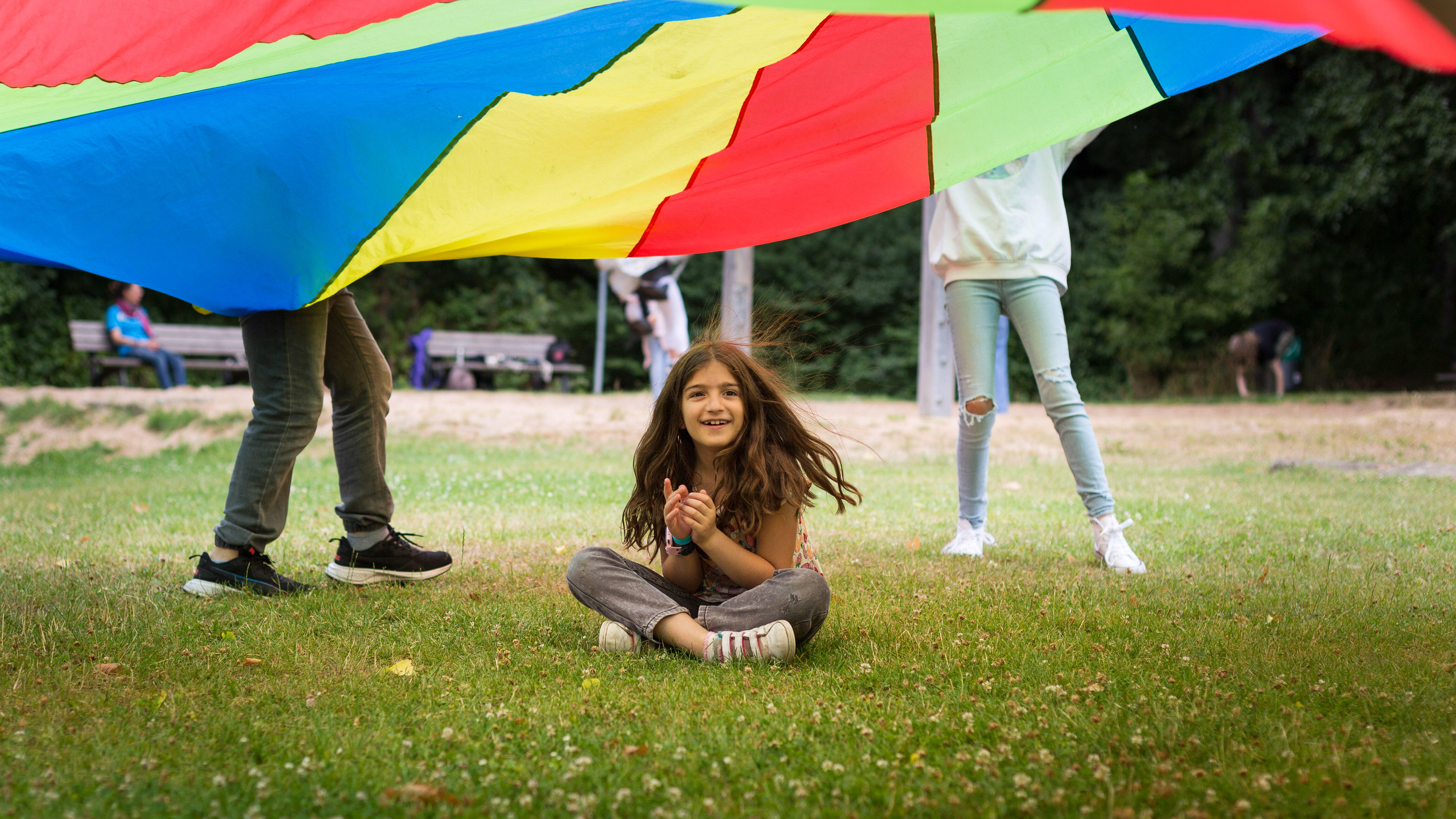 A girl sits under a colorful parachute while completing a group activity outdoors.