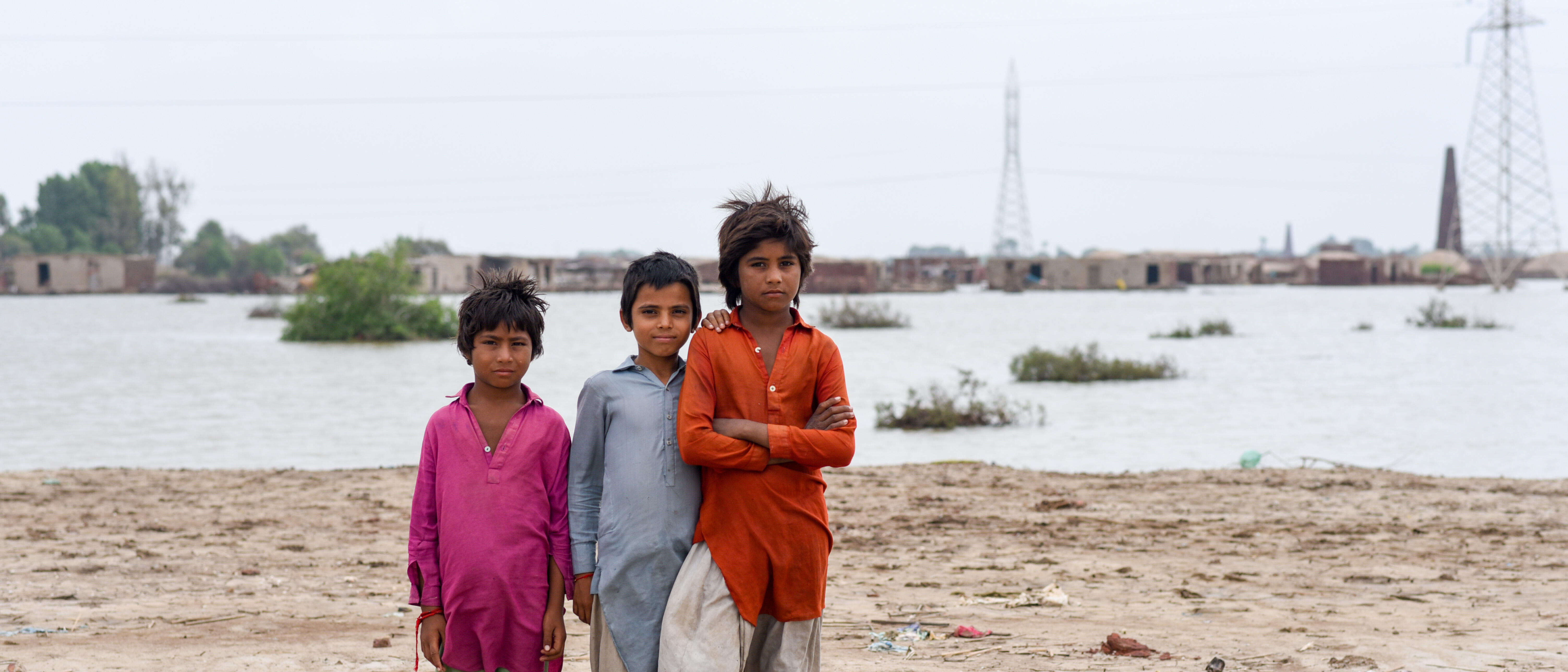 Three children pose for a photo in front of a community impacted by flooding.