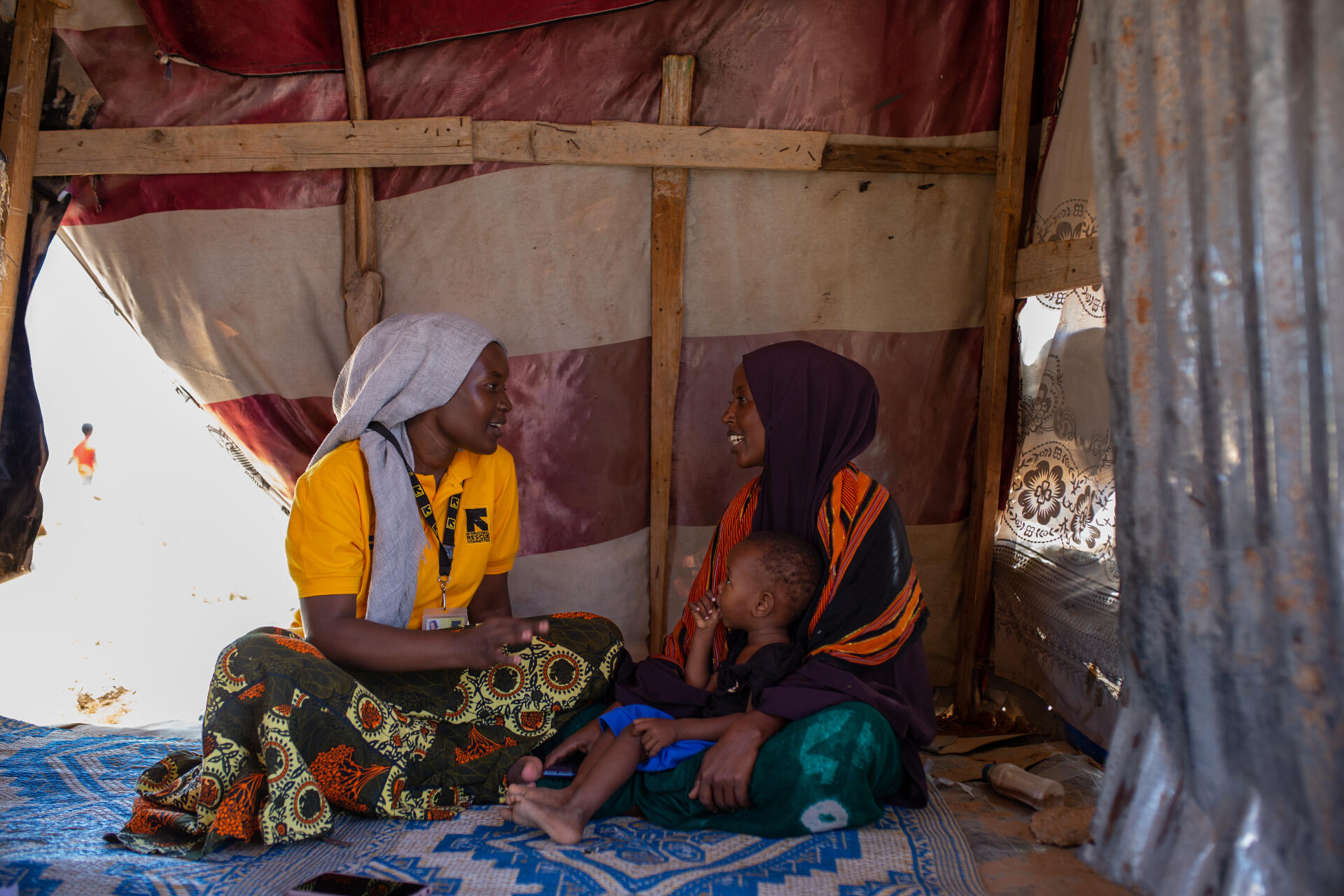 An IRC staff member consults a mother and her child inside a small building in a camp for internally displaced Somalis. The pair share a conversation while sitting on the floor.