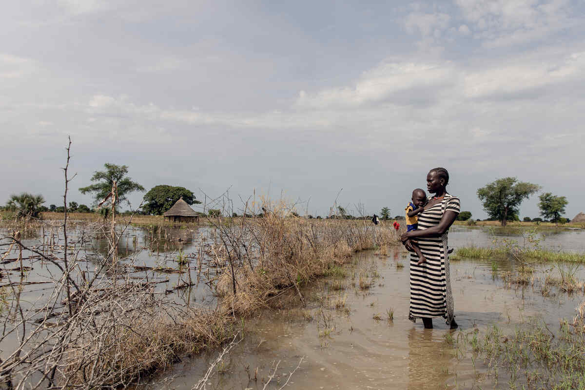 En kvinna står mitt på ett översvämmat fält och håller sitt barn i armarna.