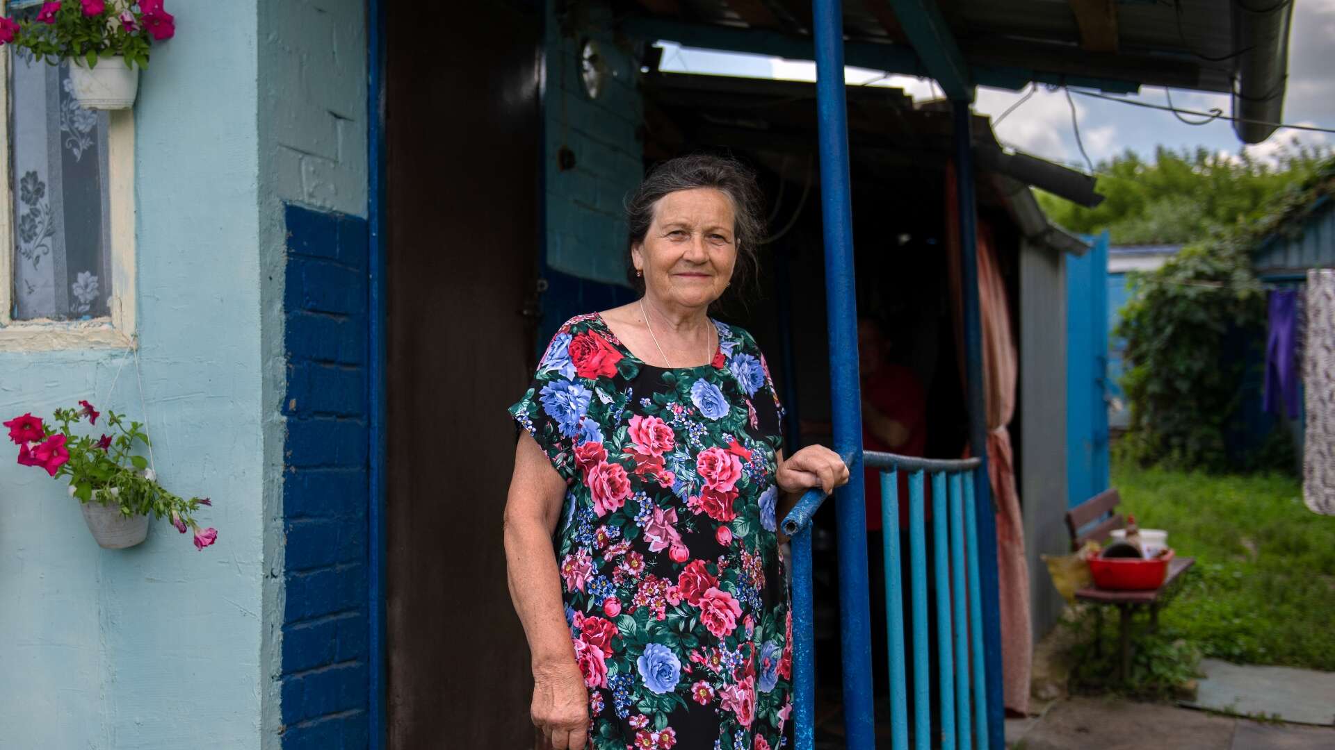 An elderly Ukrainain woman poses for a photo while standing in front of her home.