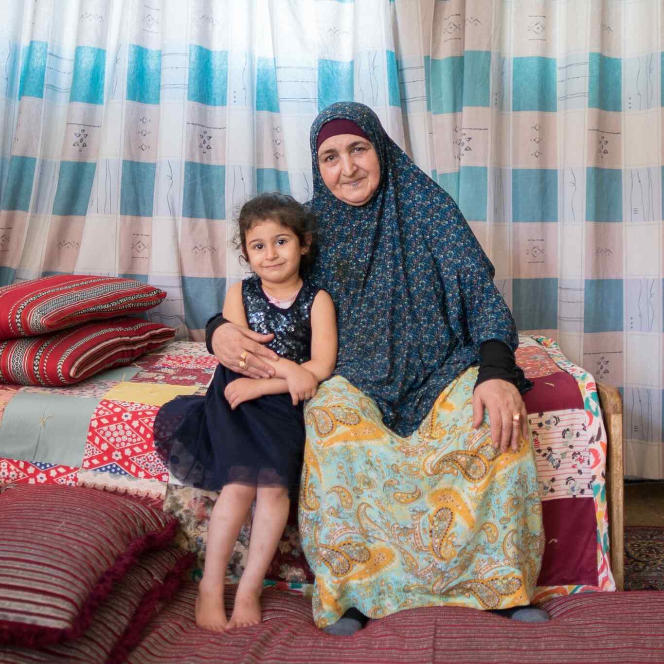 A young girl and her grandmother sit at the edge of a bed and pose for a photo.