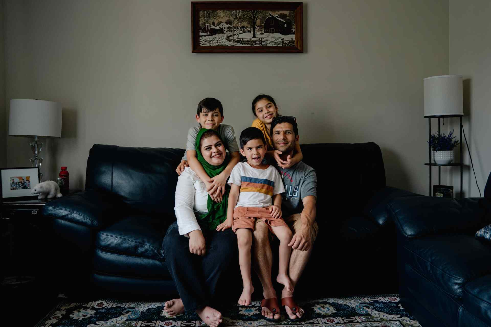 Two parents and three children pose for a portrait together on a black couch. The family was resettled after fleeing Afghanistan as refugees.
