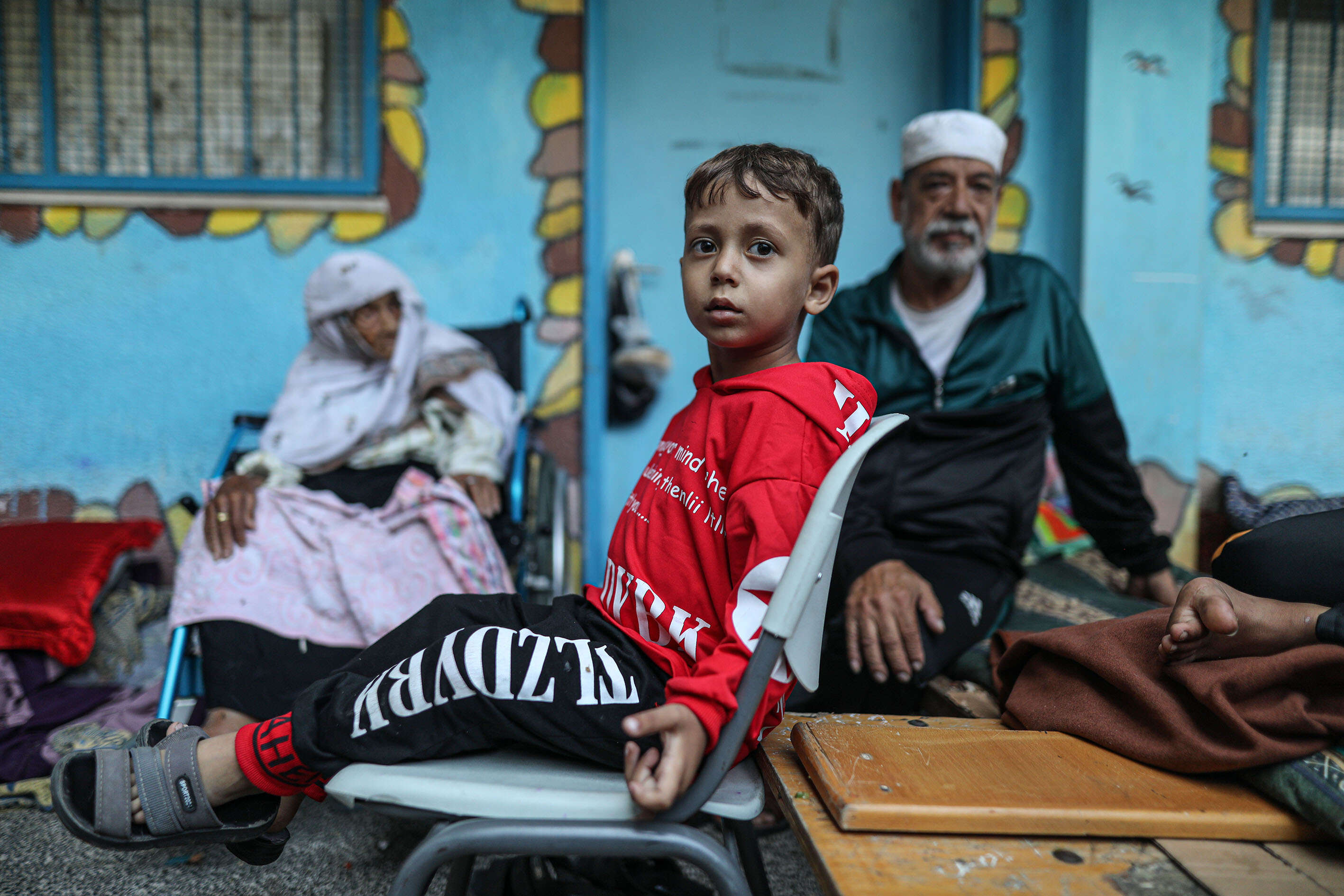 A Palestinian boy sits in a chair at a UNRWA school. An elderly woman and man sit nearby.