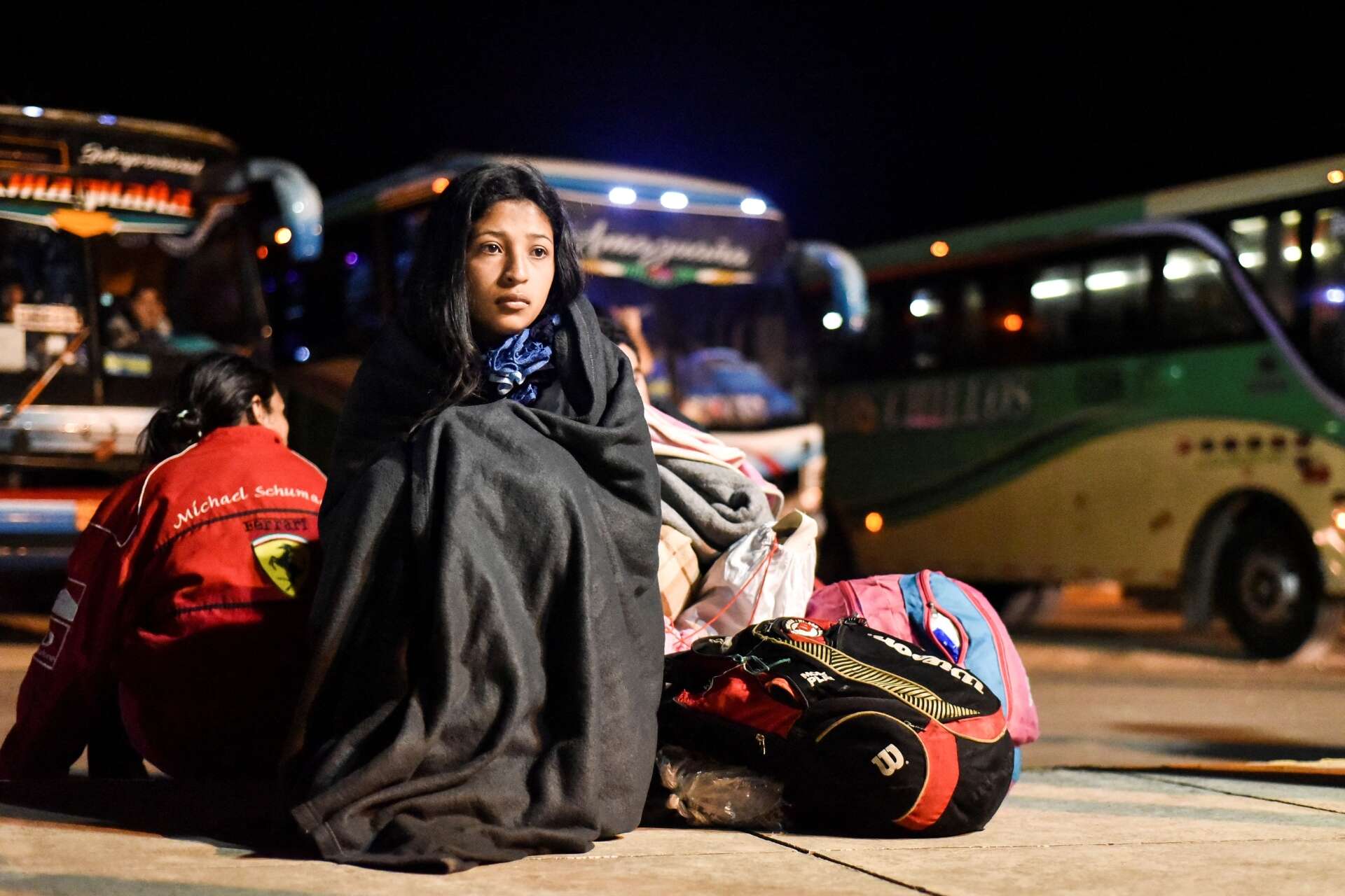 A young woman sits at a bus depot, wrapped in a blanket, next to several bags.