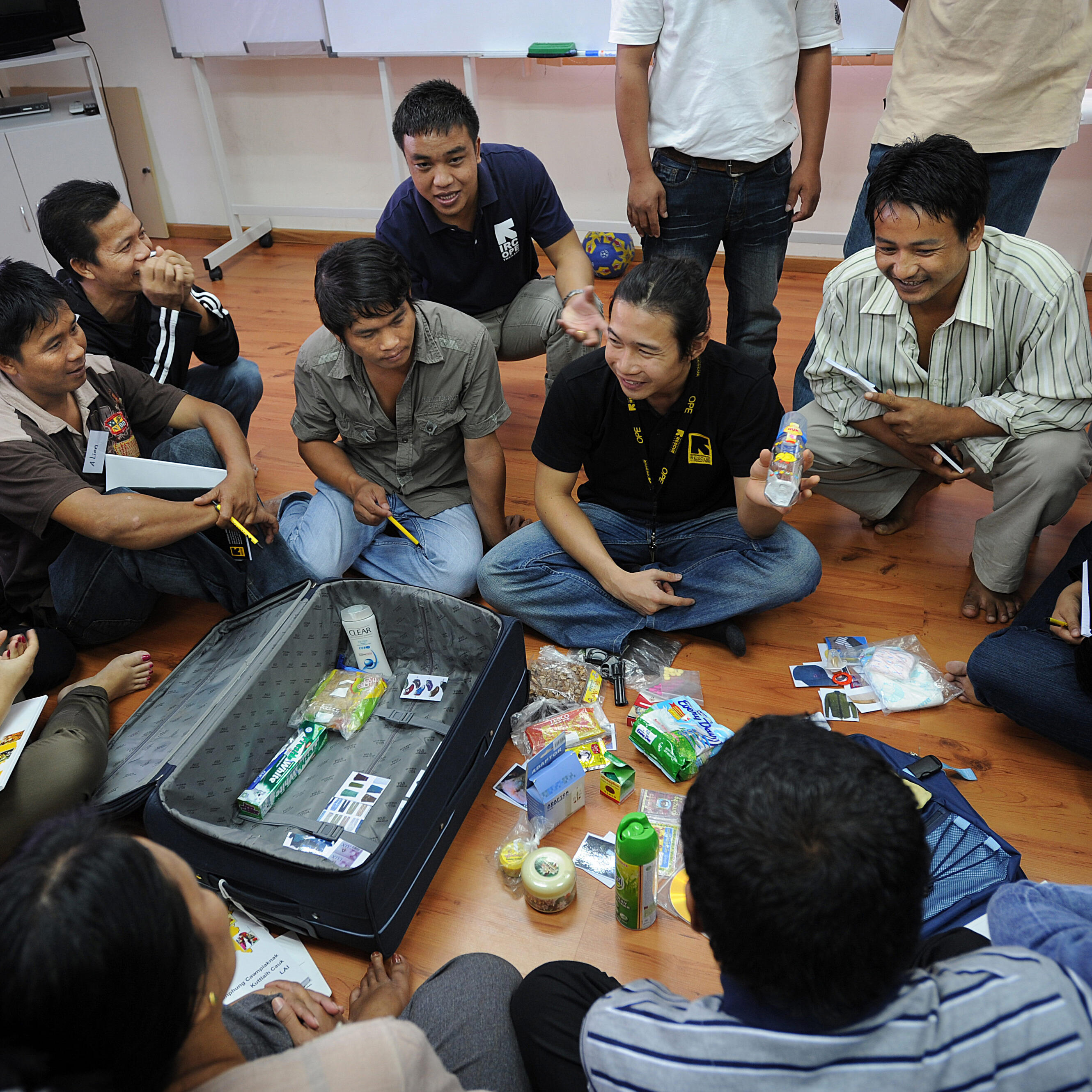 Refugees sit on the floor around an International Rescue Committee volunteer and suitcase as they learn how to strategically pack luggage.