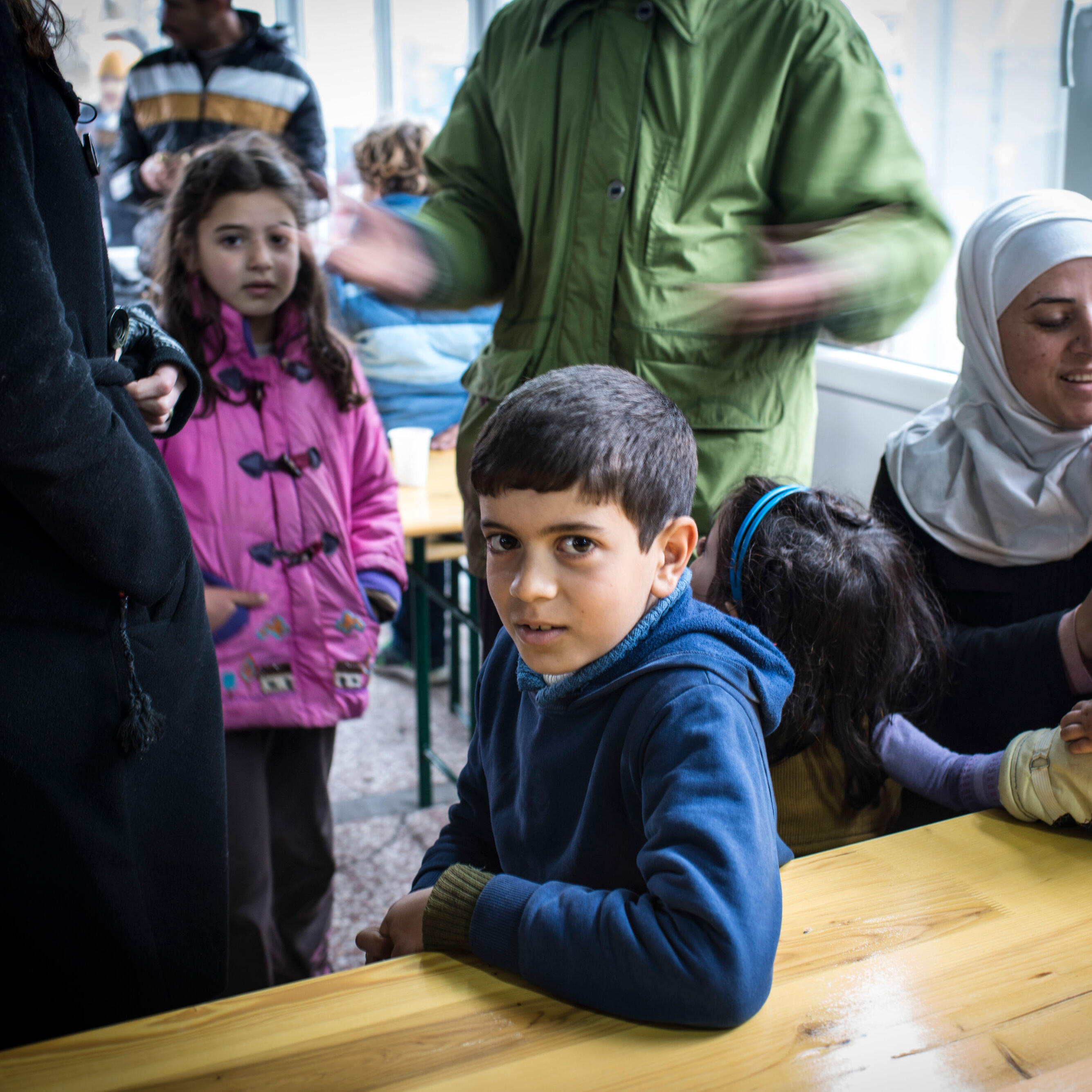 A young boy sits at a table in a refugee shelter in Serbia, surrounded by other children and their families seeking safety in Europe.