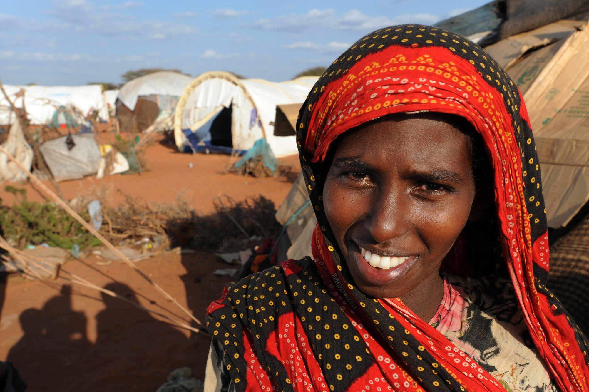 A smiling young woman stands outside a tent in Dadaab refugee camp in Kenya on a sunny day,