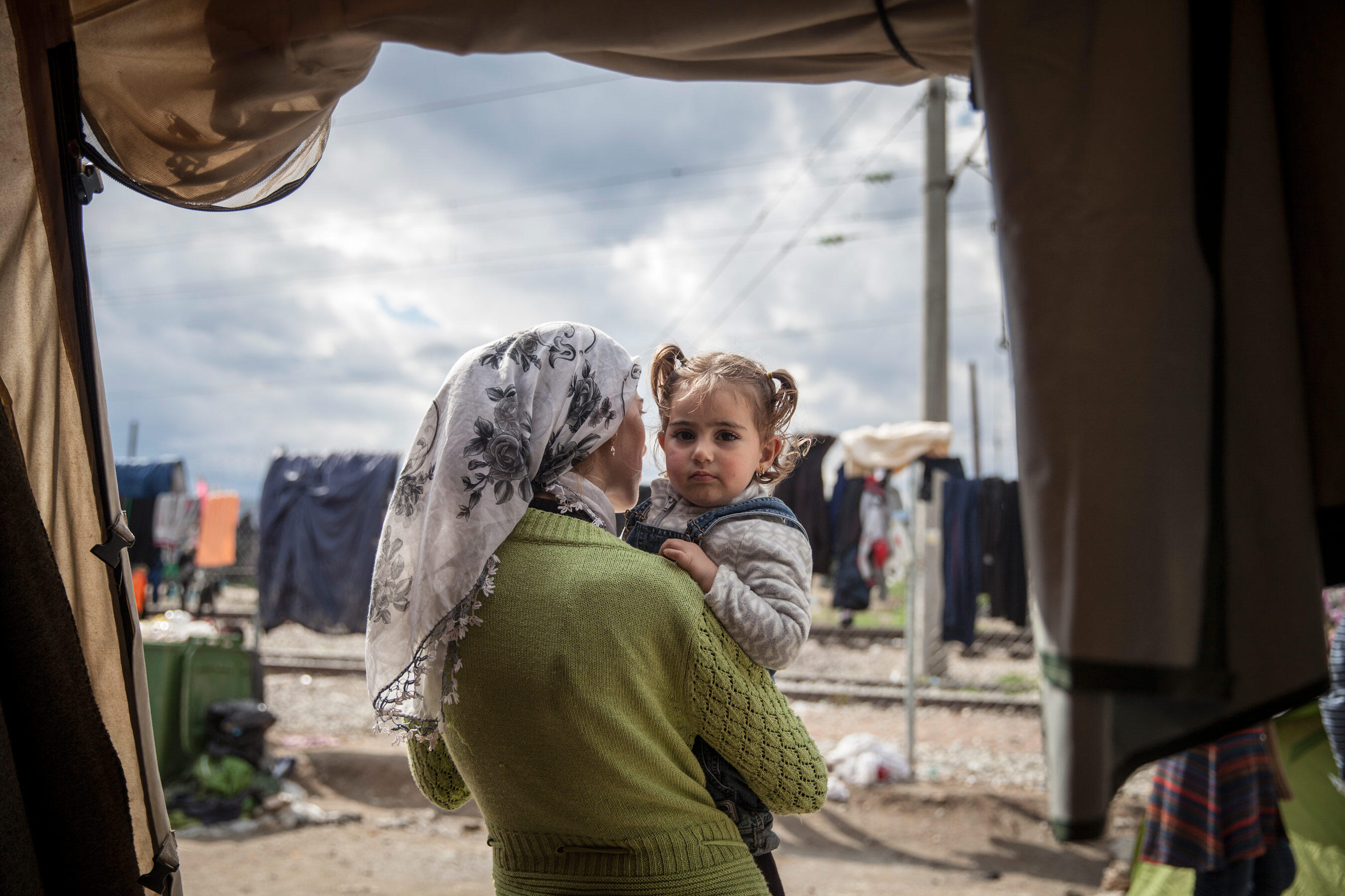 A mother holds her toddler daughter and looks out from their tent on a cloudy day in a refugee camp in Idomeni, Greece.