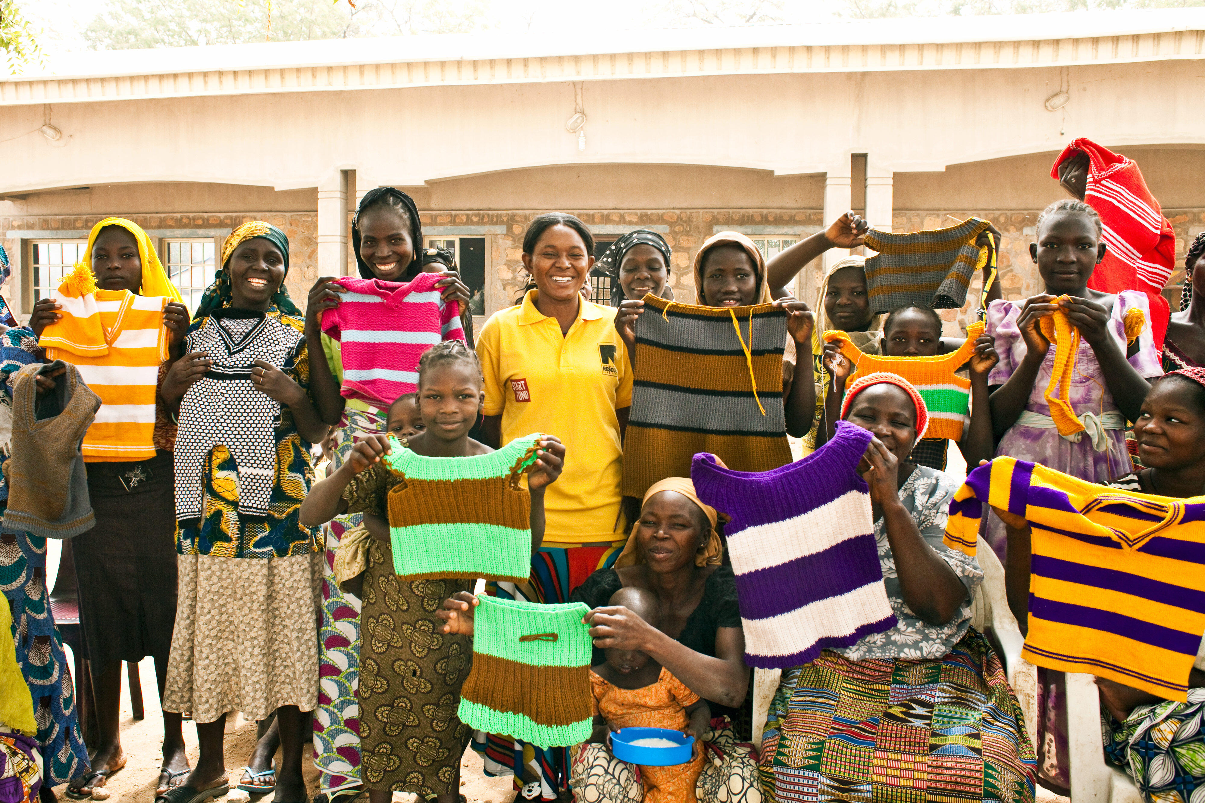 A group of women who are part of an IRC livelihoods program in Nigeria stand outside, lifting hand-knitted tops to show them off.  