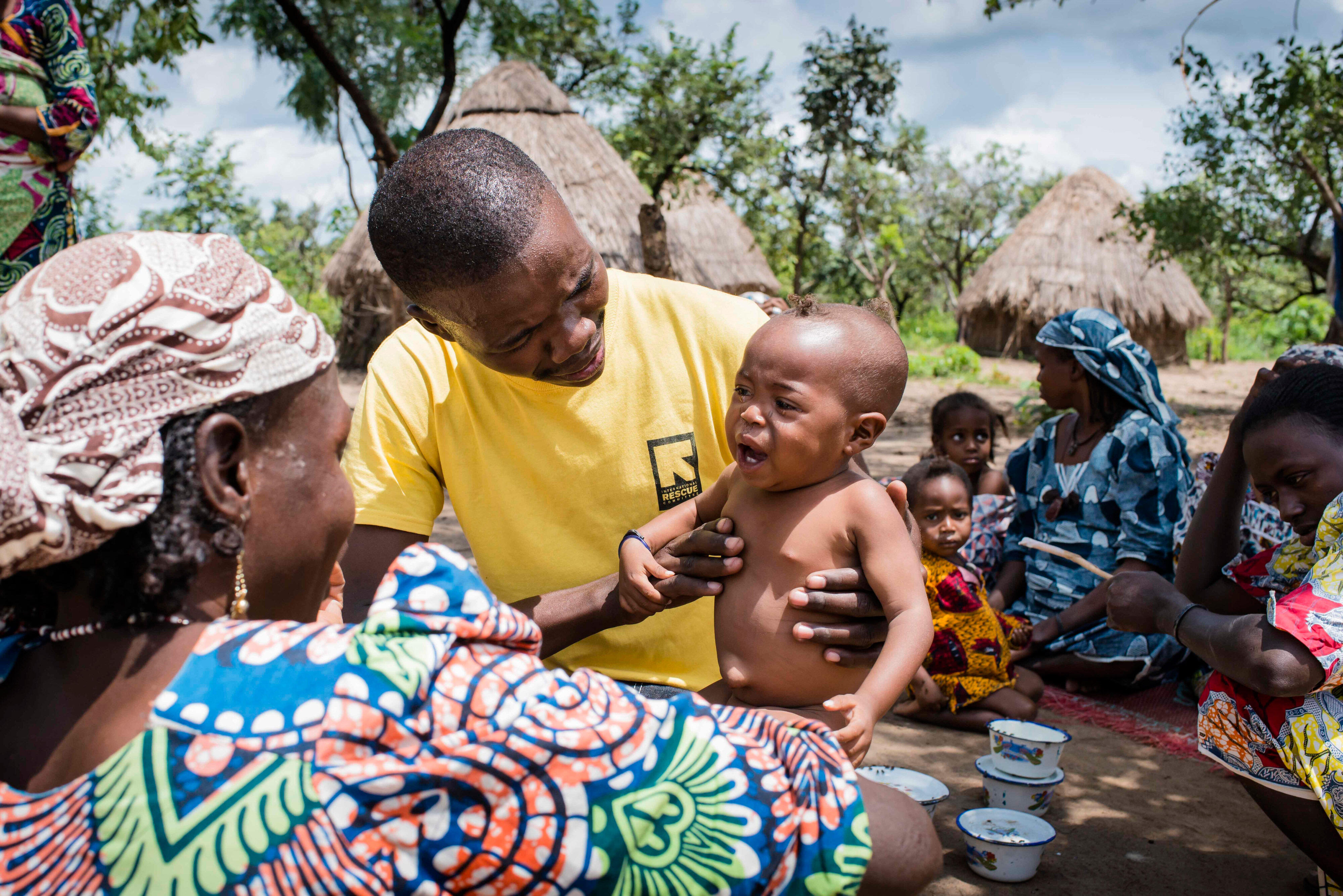 An IRC health worker holds a crying baby, giving him a medical checkup in a village in Central African Republic as his mother looks on smiling. 