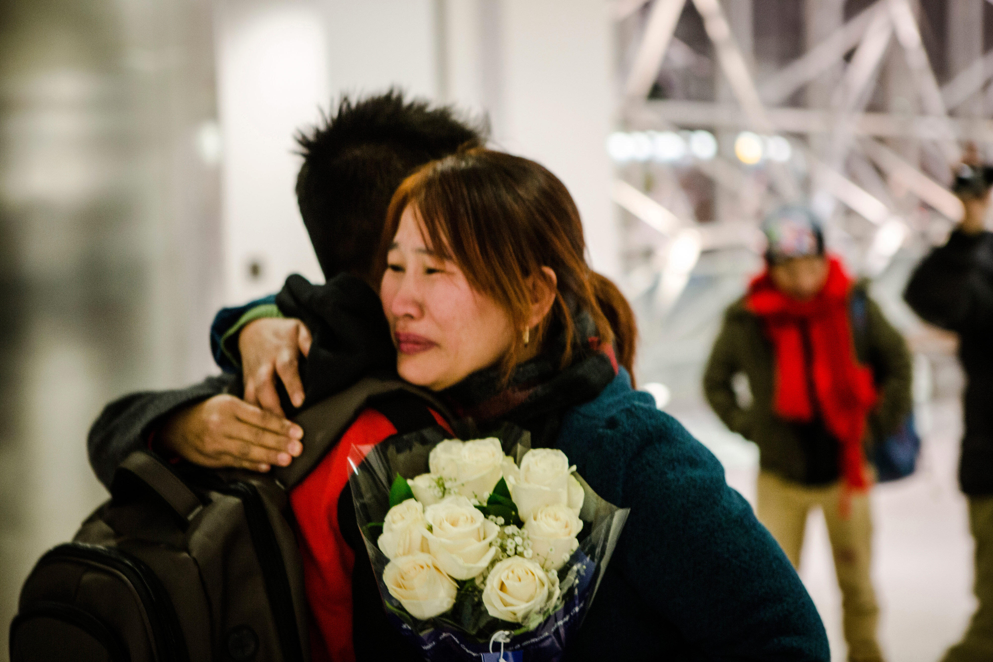 Crying mother hugs son while holding flowers at the airport.