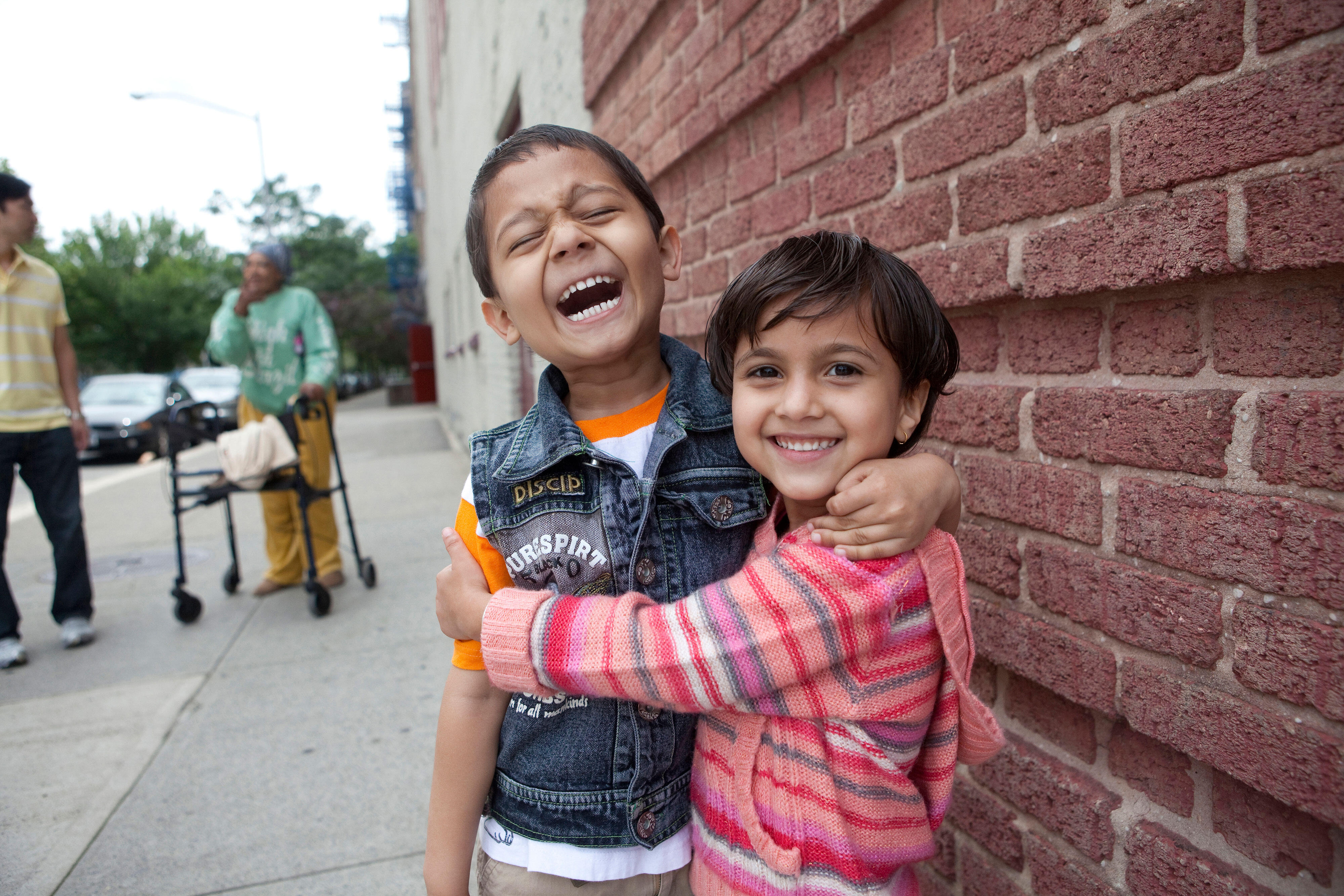 A young boy and girl stand outside in front of a brick wall, laughing and embracing.