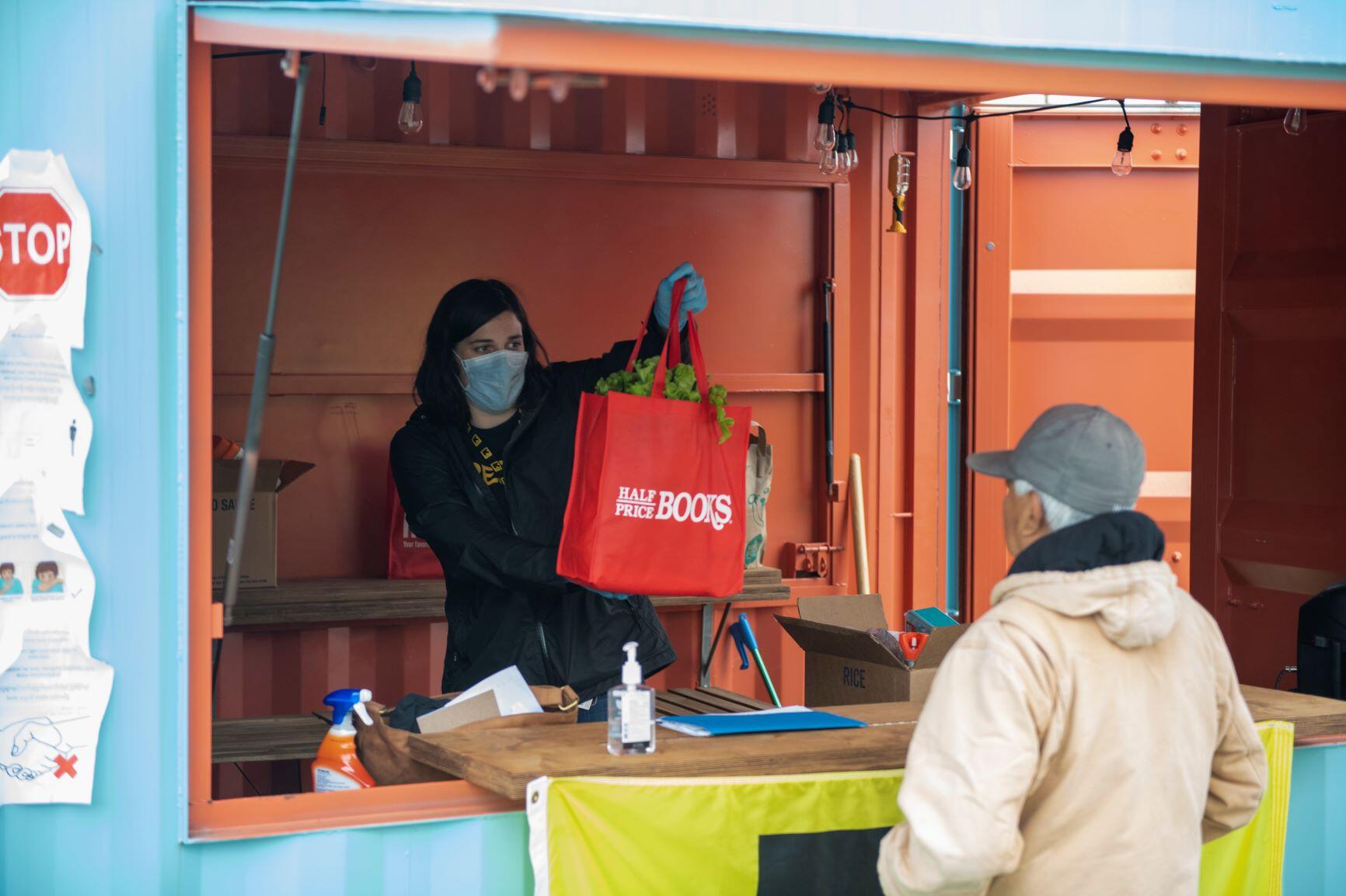 Isabelle Chamberlain, wearing a mask, holds up a red bag full of food that she is about to hand to an IRC client. 