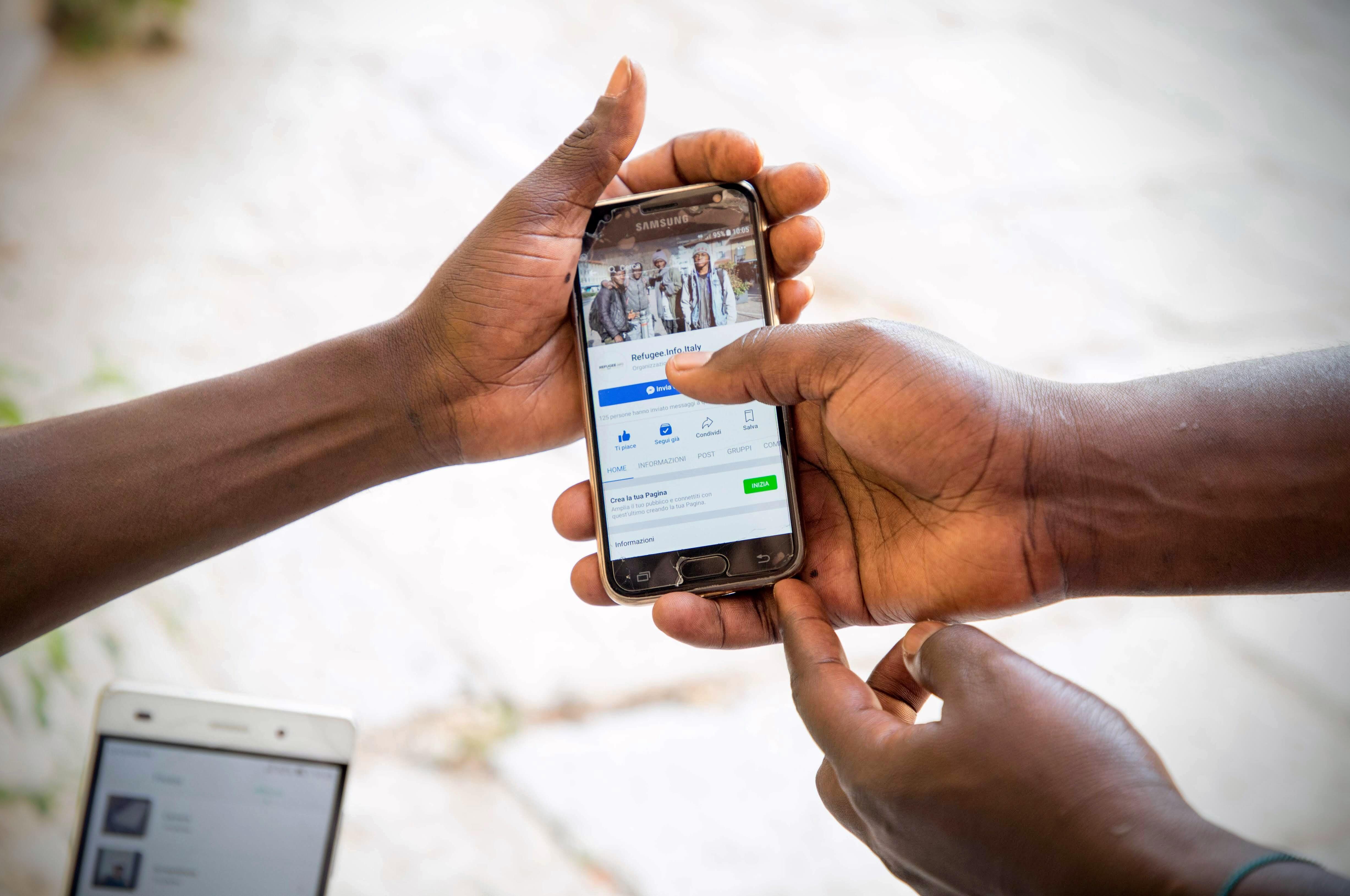 Hands holding a cellphone displaying an IRC Refugee.Info Facebook page on-screen