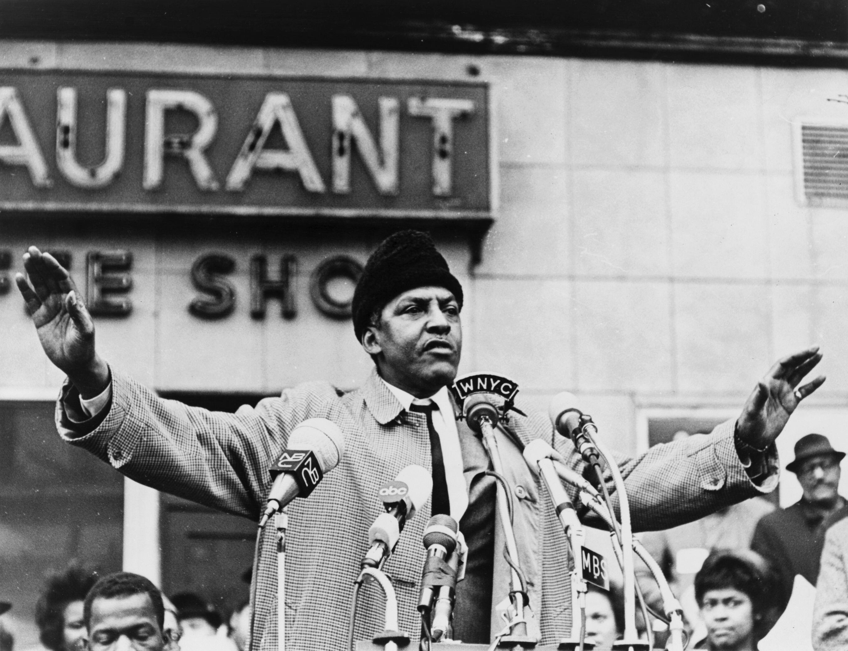 Bayard Rustin stands before microphones at a podium with arms outstretched