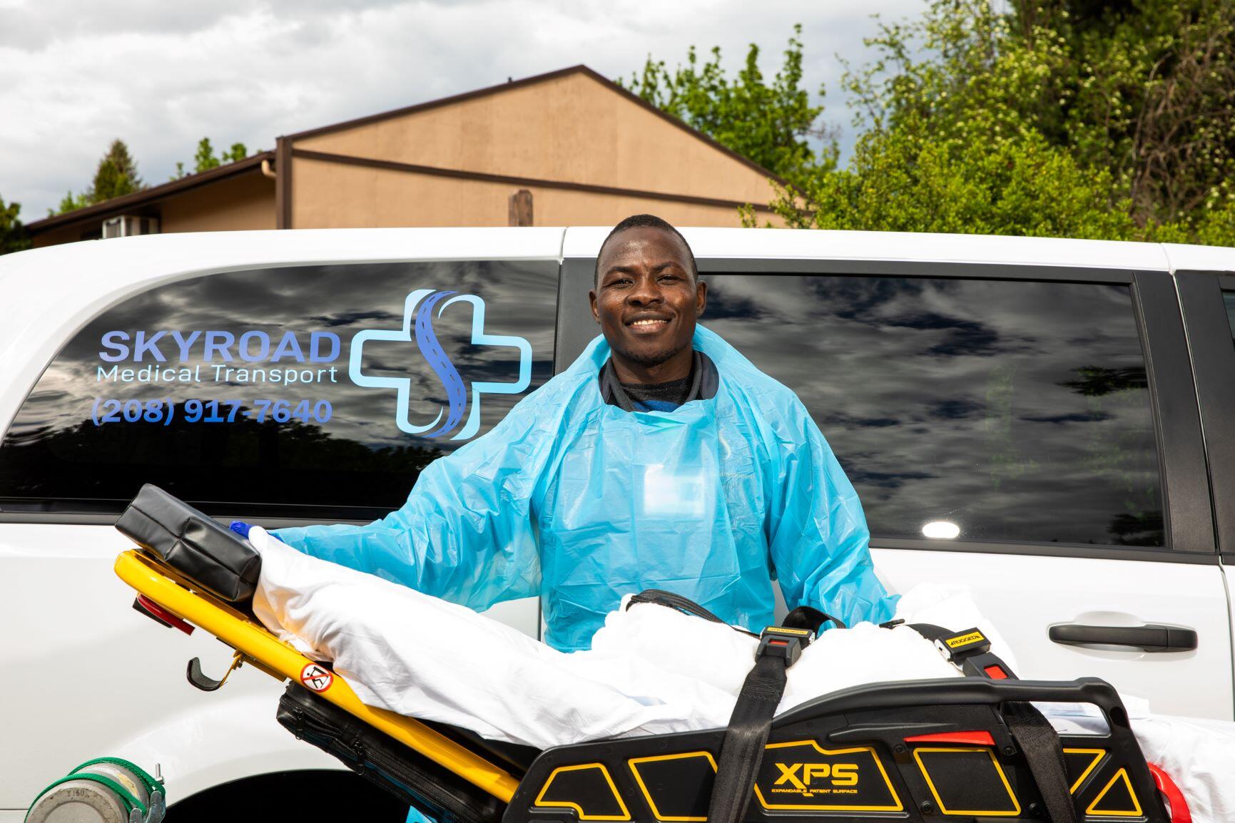 Jonathan Amissa, who owns a medical transport business, poses with a stretcher in front of one of his vans. He is wearing bright blue scrubs. 