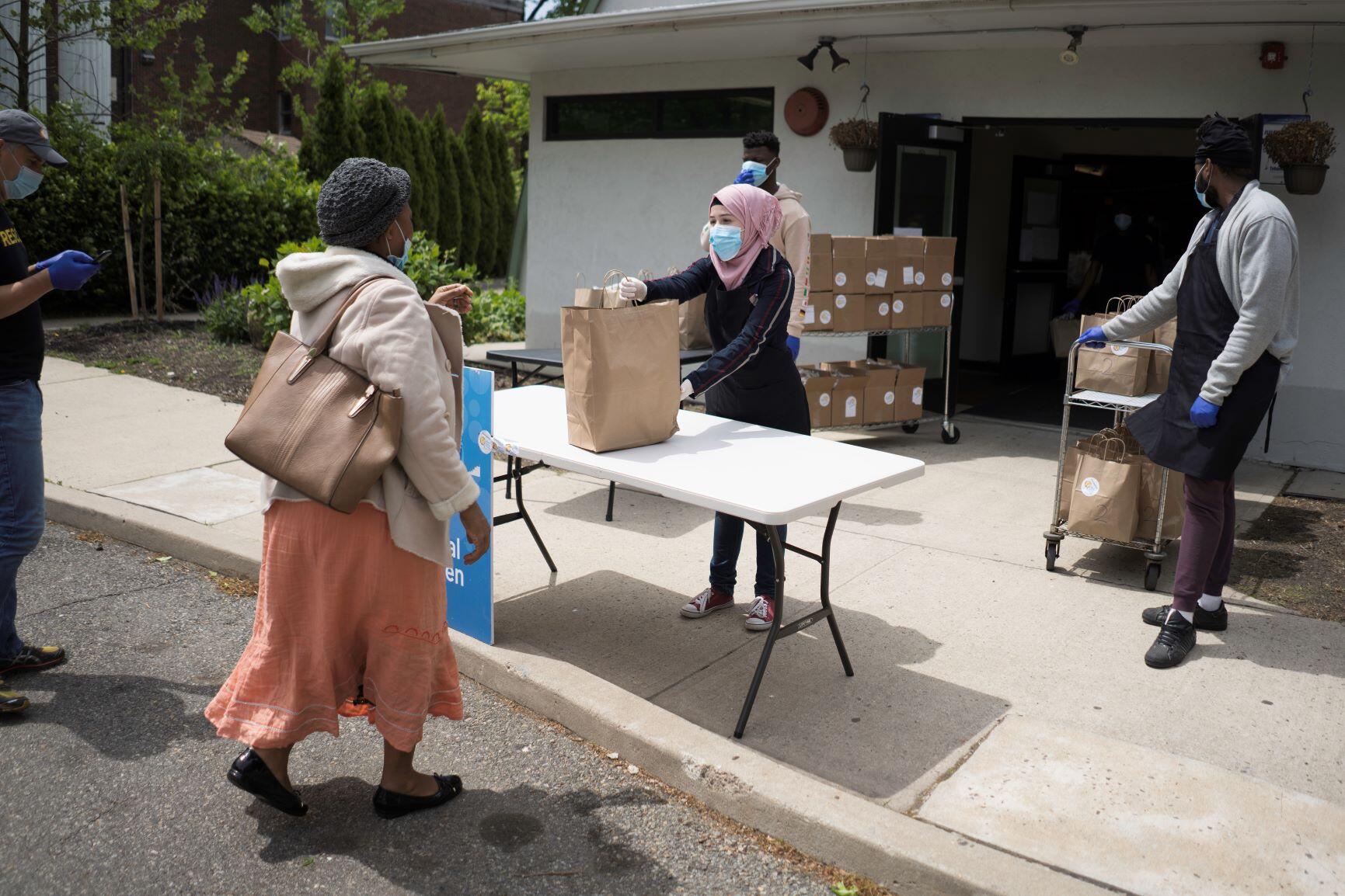 Rania Abou, a nineteen-year-old refugee from Syria, hands out food at her job with WC Kitchen and the IRC in Elizabeth, New Jersey. She is wearing a mask, a pink head scarf, and a black jacket and is handing a paper grocery bag to a woman in a pink skirt.