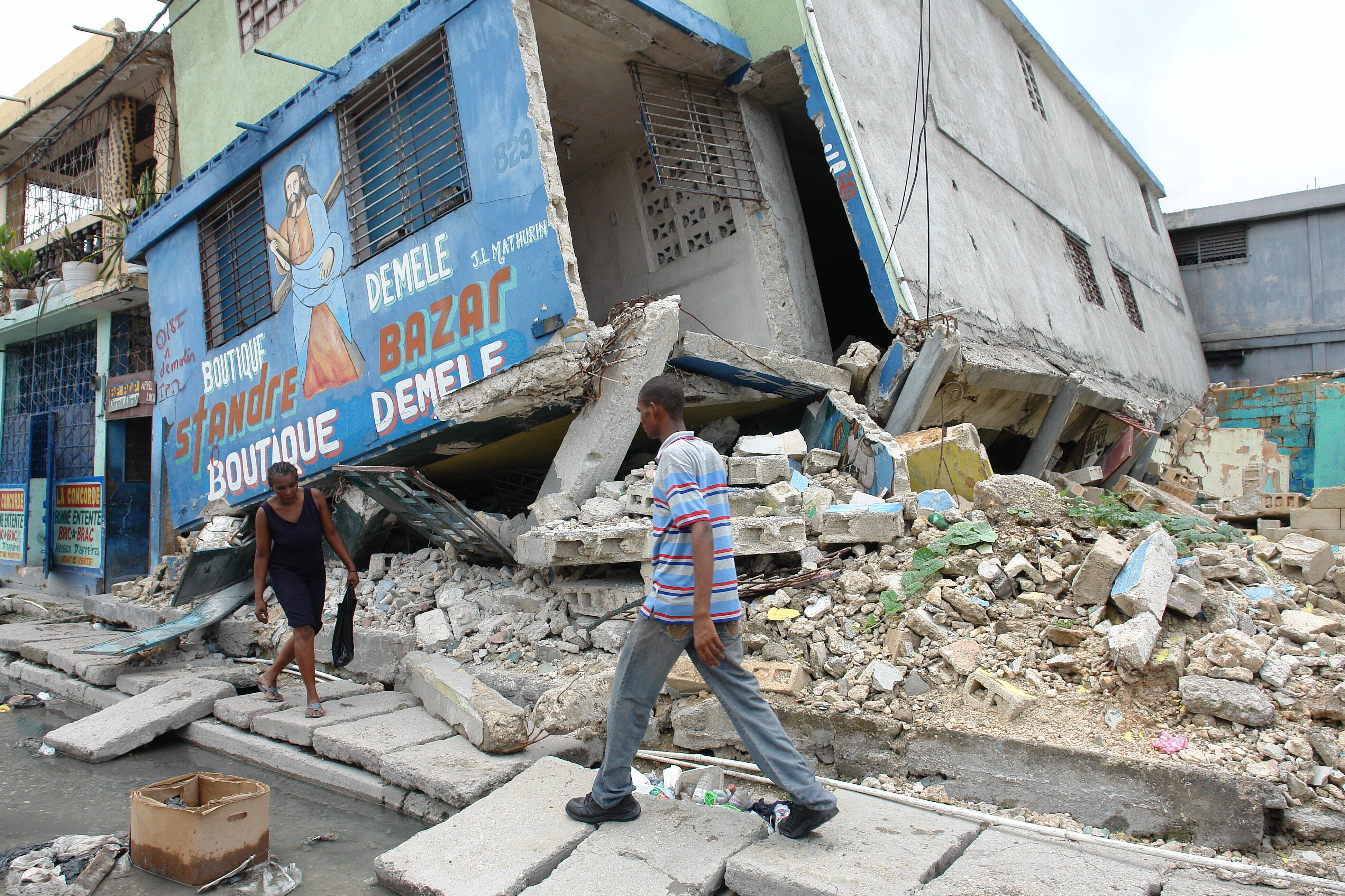 A man and a woman walk carefully on a damaged sidewalk past collapsed buildings in Port-au-Prince, Haiti after the 2010 earthquake.