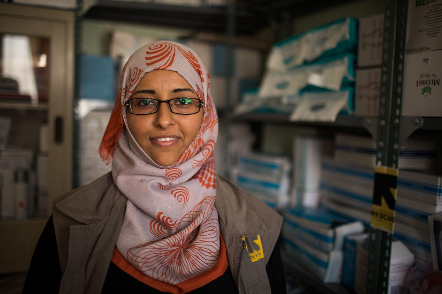 Dr. Rasha Rashed, a doctor and reproductive health manager for the IRC in Yemen, stands in a room with media supplies. She is wearing a red and white scarf and an IRC-branded vest. 