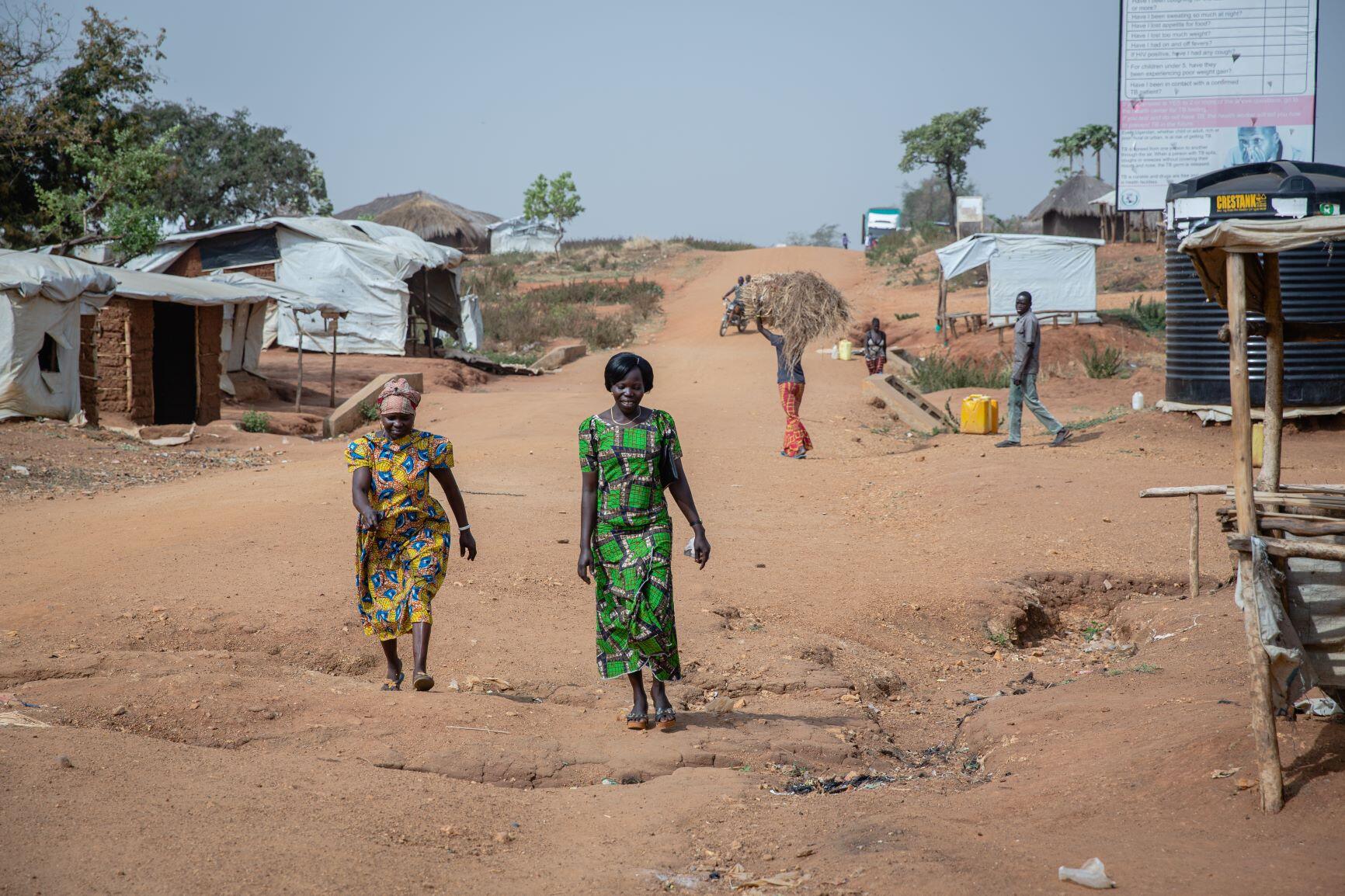 Women's rights activists Foni Grace and Loyce Tabu walk in Bidi Bidi refugeee settlement in Uganda.They are at the forefront of the photo and there are small makeshift homes and a man carrying a bale of hay behind them. 