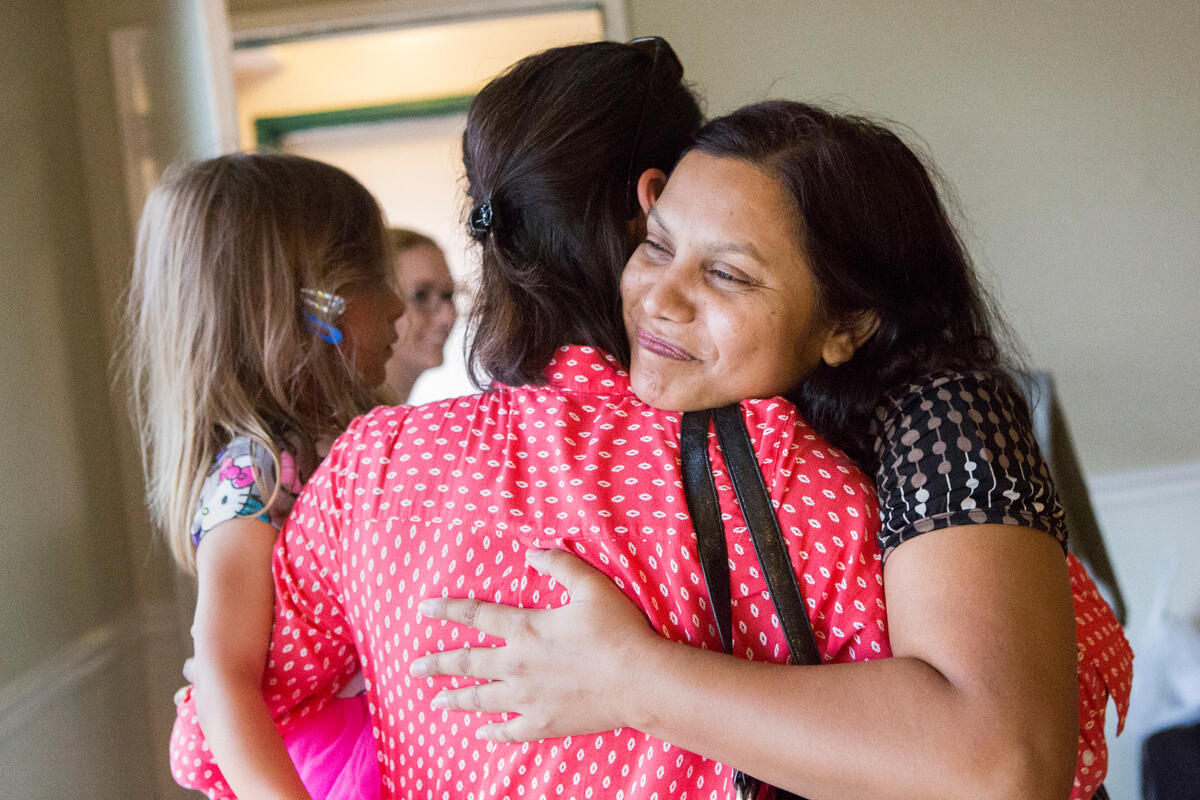 Two women hug while one holds a small child. We see one of their faces, she is smiling.