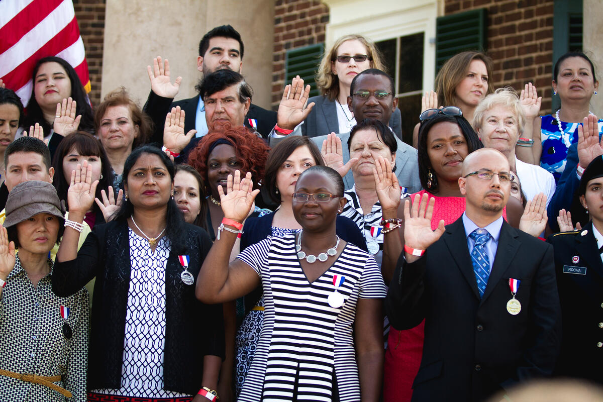 Two new citizens take their oath. They are standing in a large group of people and holding up their right hands. 