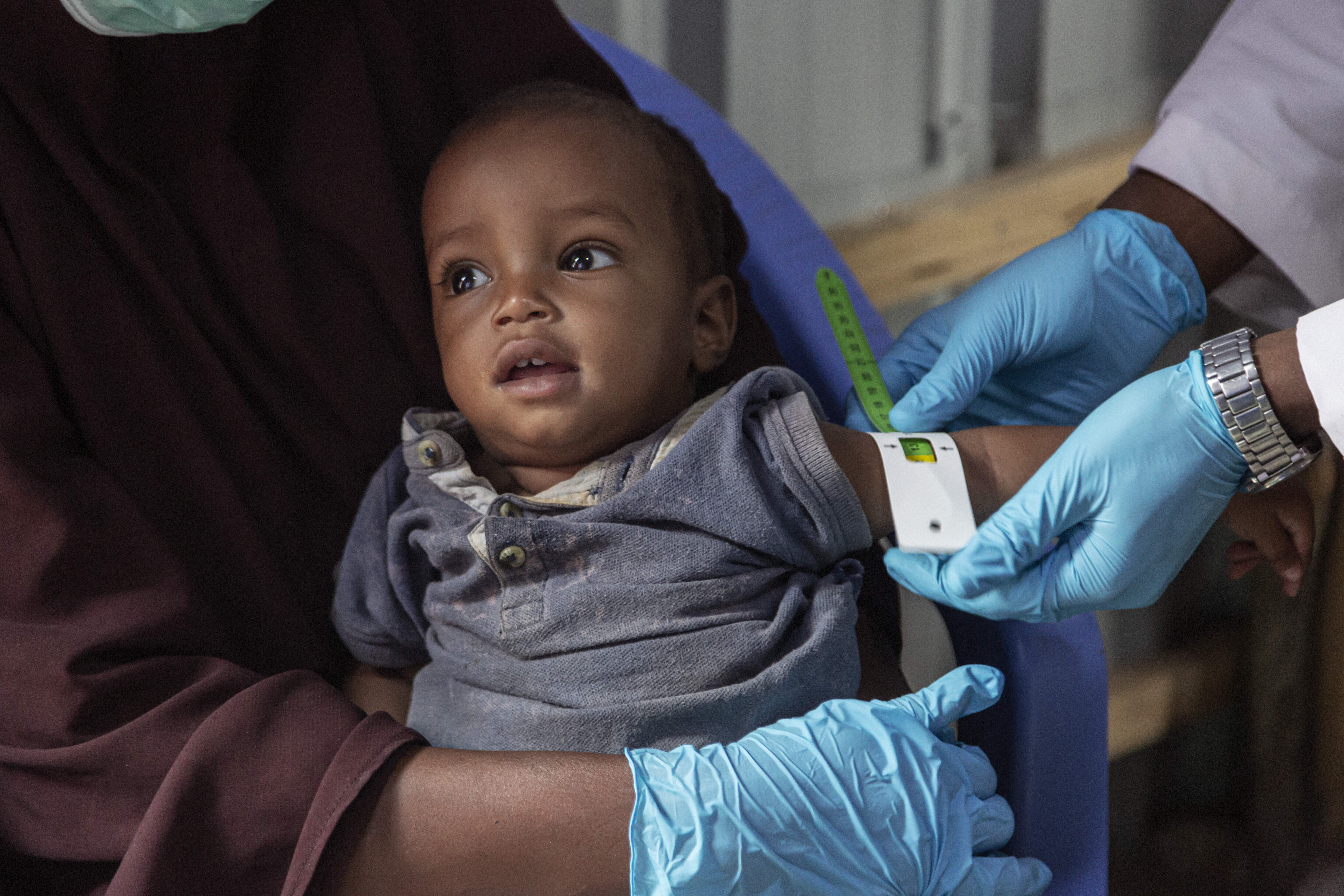 Amina's son, while sitting on his mother's lap, has his arm circumference measured by an IRC staff member wearing blue gloves. 