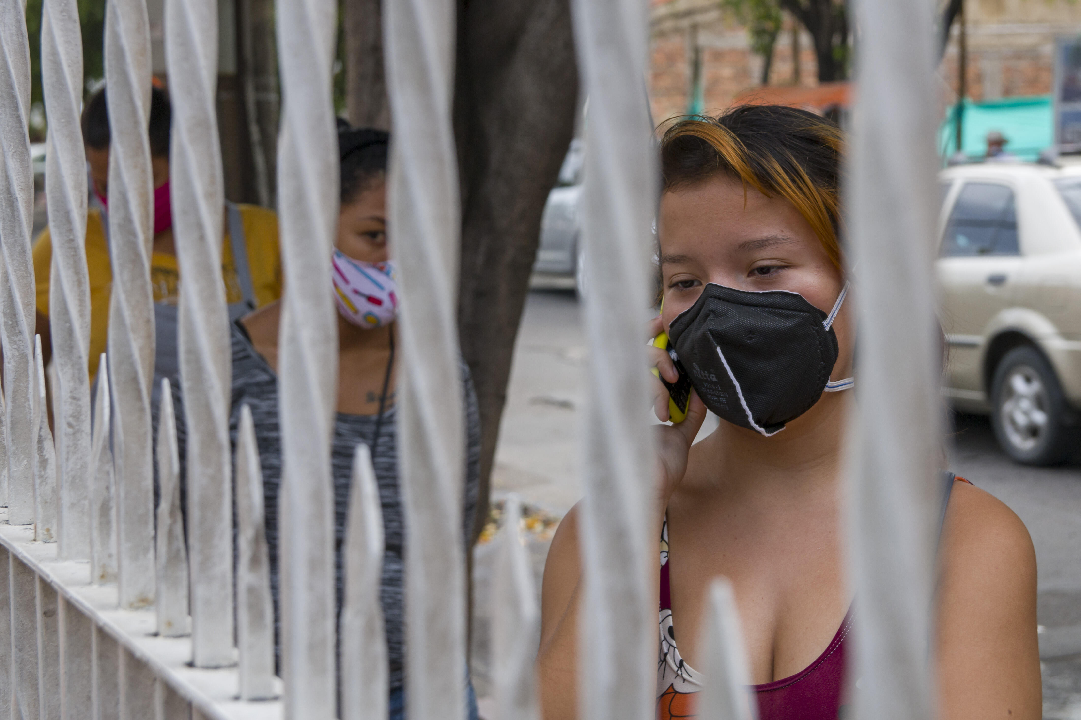 Women wearing face masks wait alongside a fence to enter an IRC health clinic on the Colombia/Venezuela border. One talks on her mobile phone.
