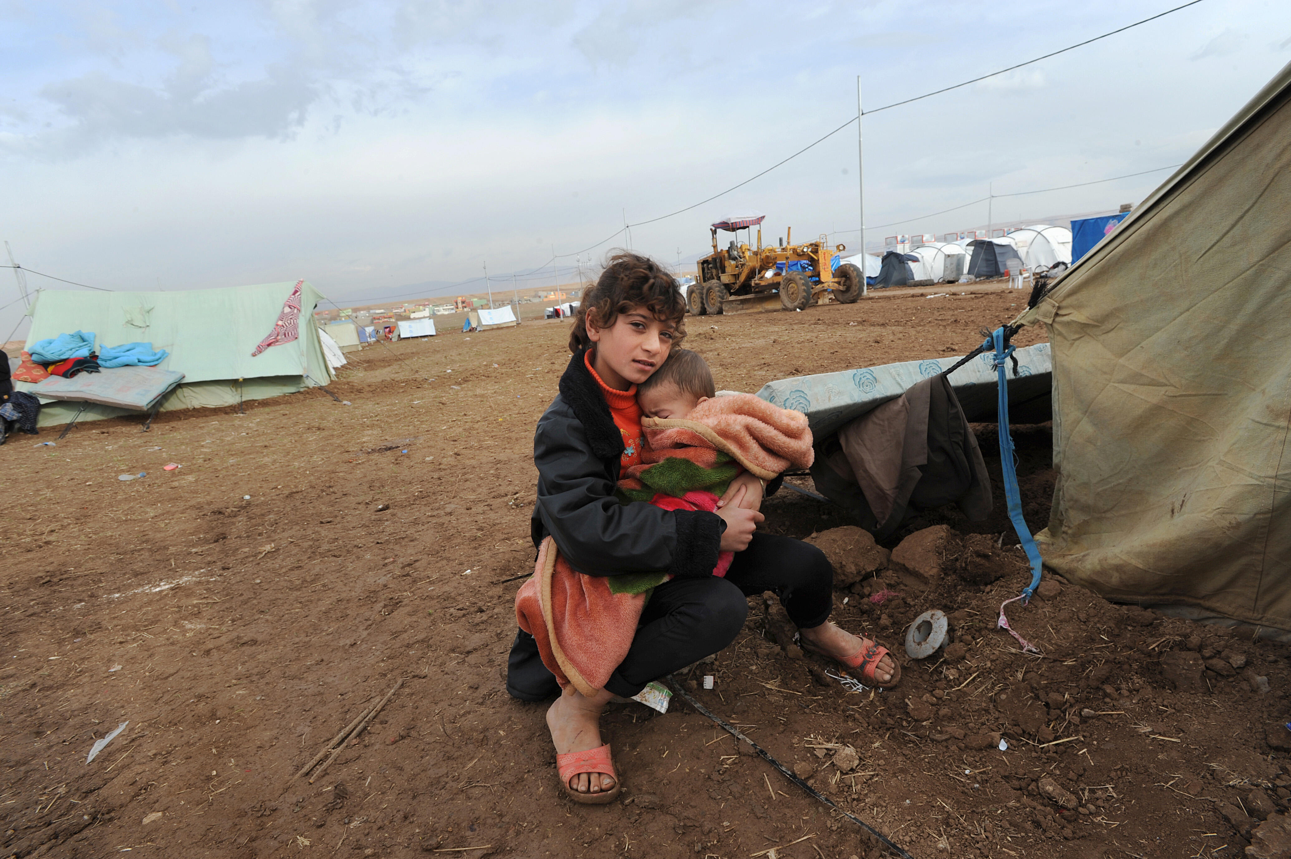 A displaced girl sits just outside a tent in Dohuk, Iraq holding a young child in her arms, bundled in a blanket.