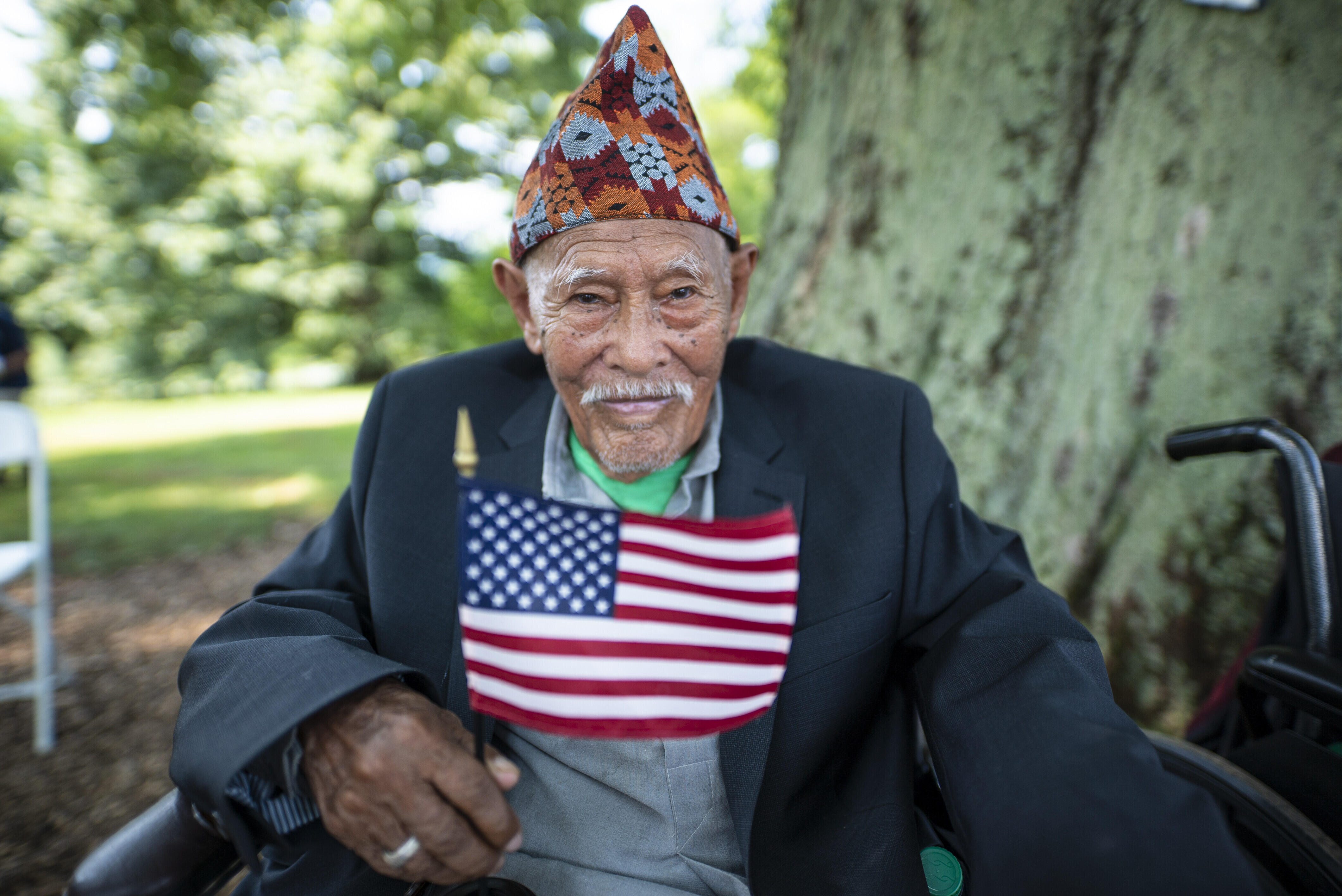 An older man and refugee at a citizenship ceremony sits in front of a tree holding an American flag