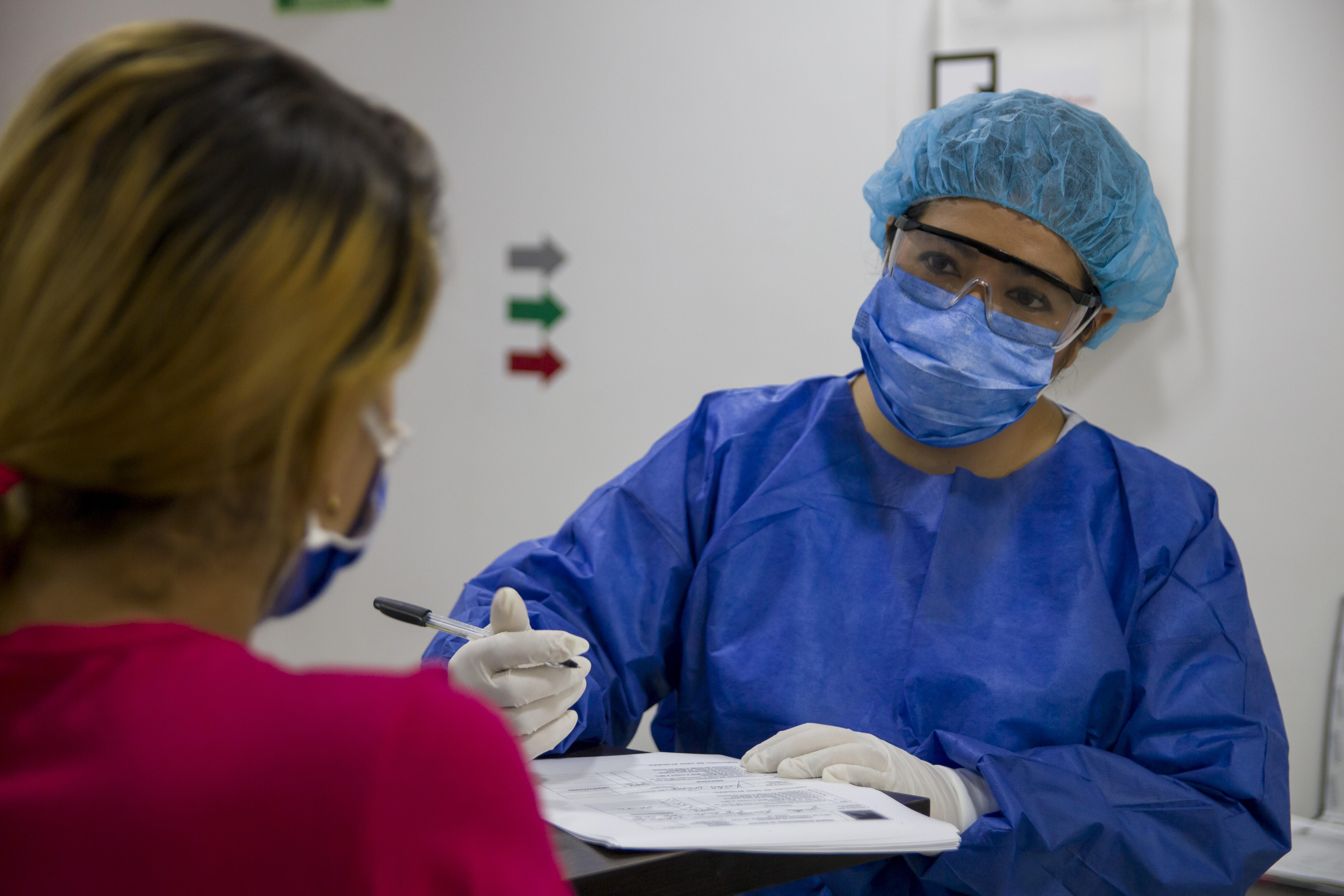 An IRC health worker wearing PPE to protect against COVID-19 holds a pen as she speaks with a patient to complete her paperwork.