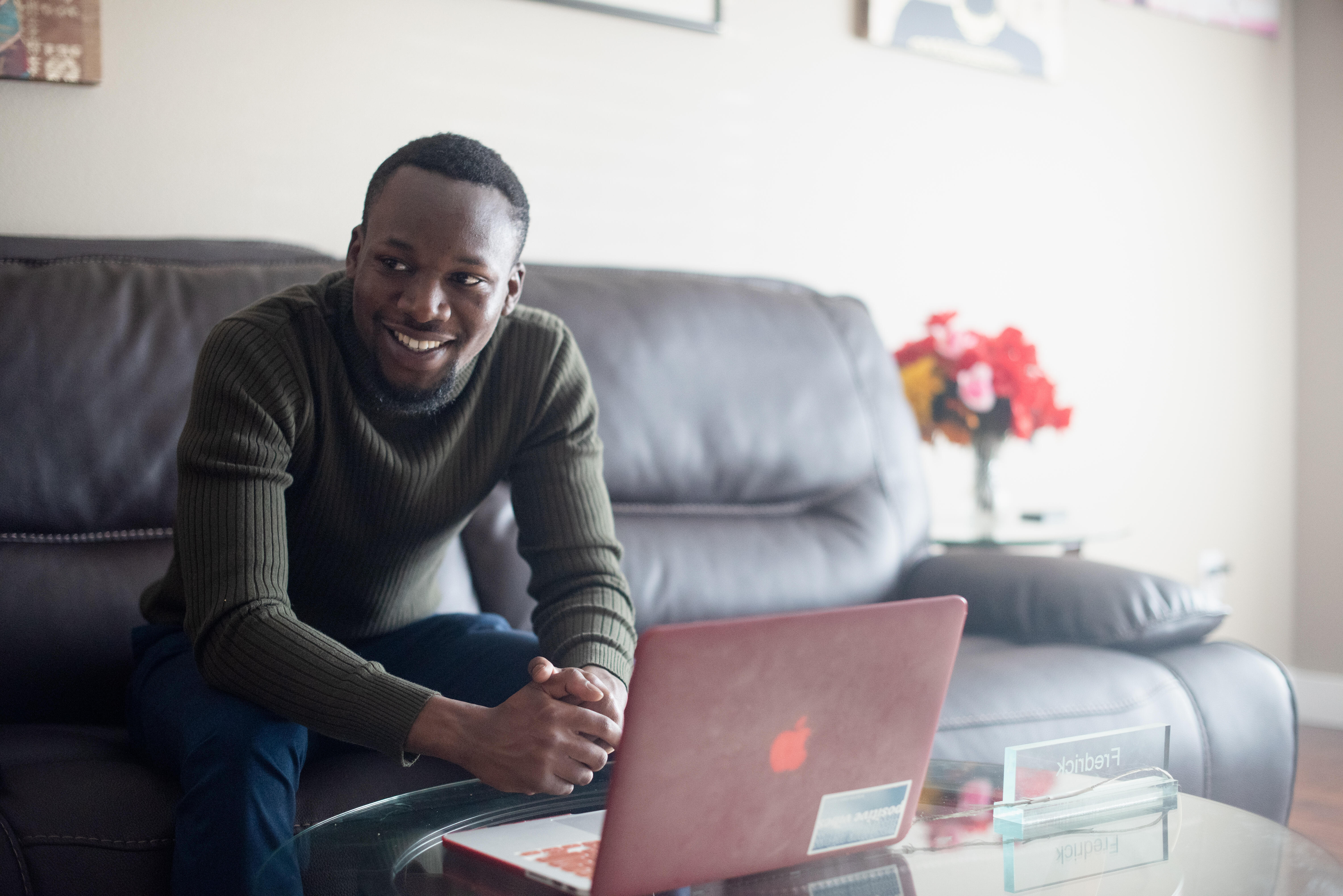 Fredrick sits on a couch in his living room with a laptop on a table in front of him. He is looking at the camera and smiling. 