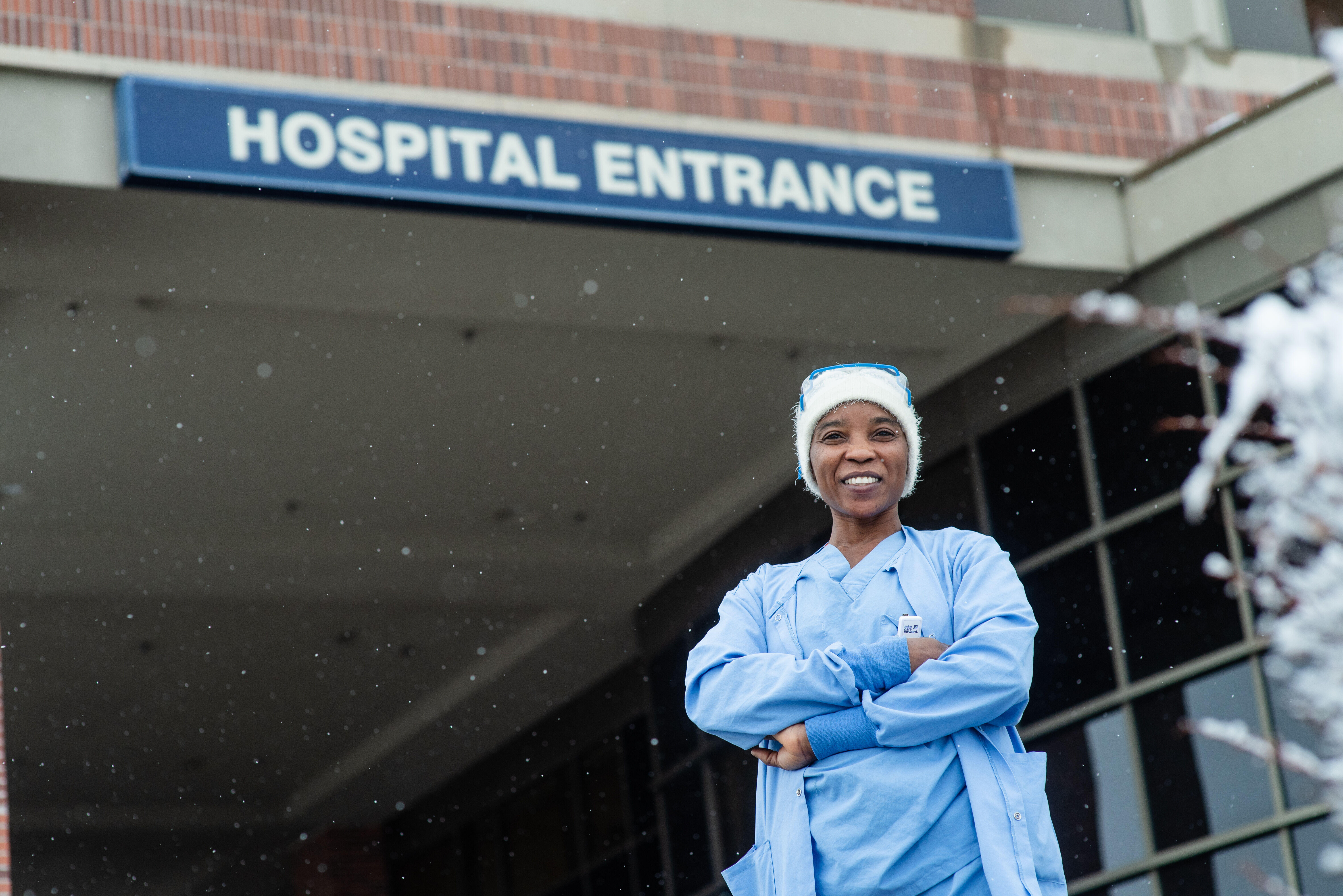 Nabila stands with her arms crossed and smiling in front of the hospital where she works. She is wearing blue scrubs and a white winter hat.