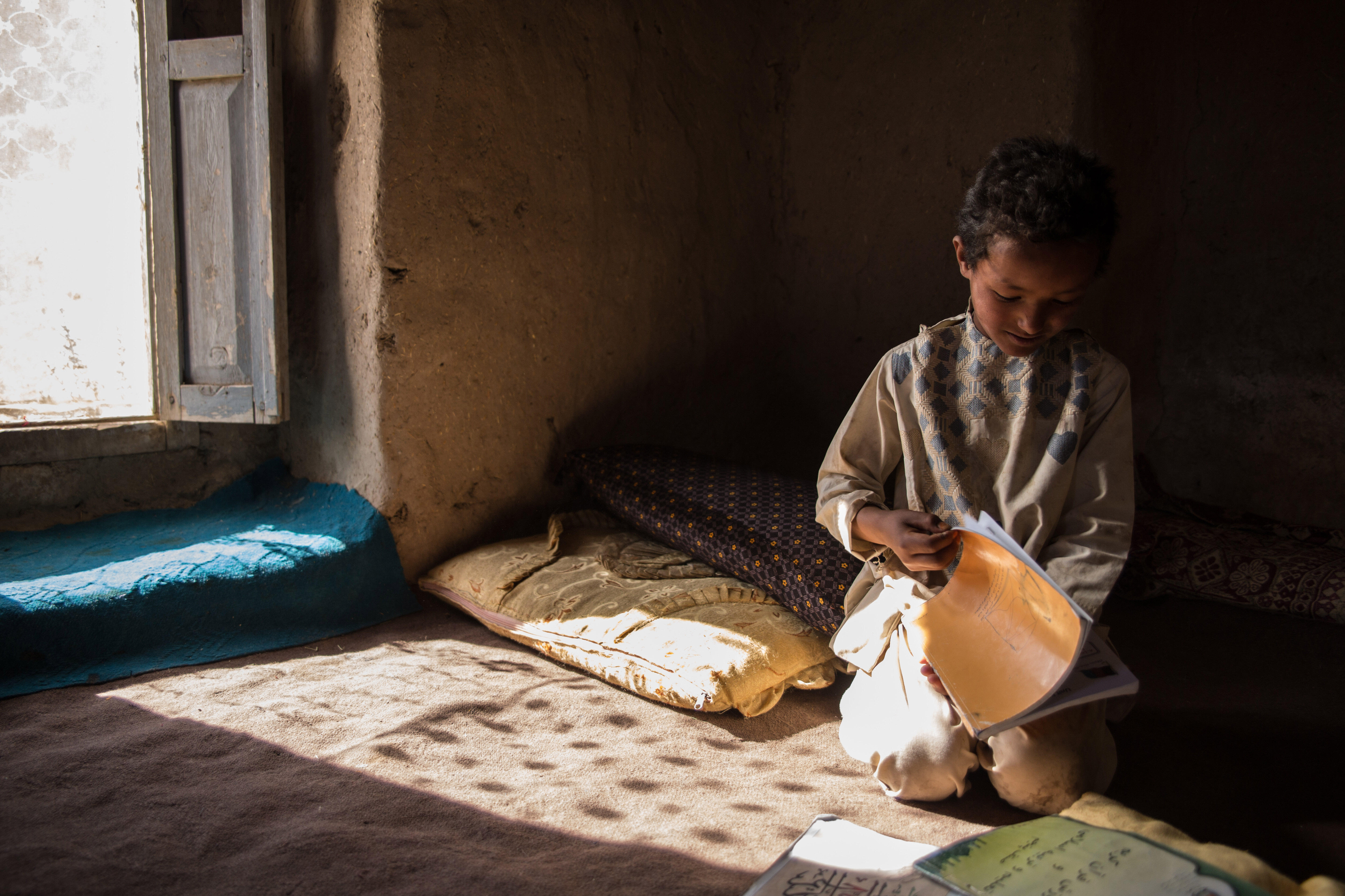 A young boy sits on a mat reading a school exercise book in his family's home in Afghanistan.