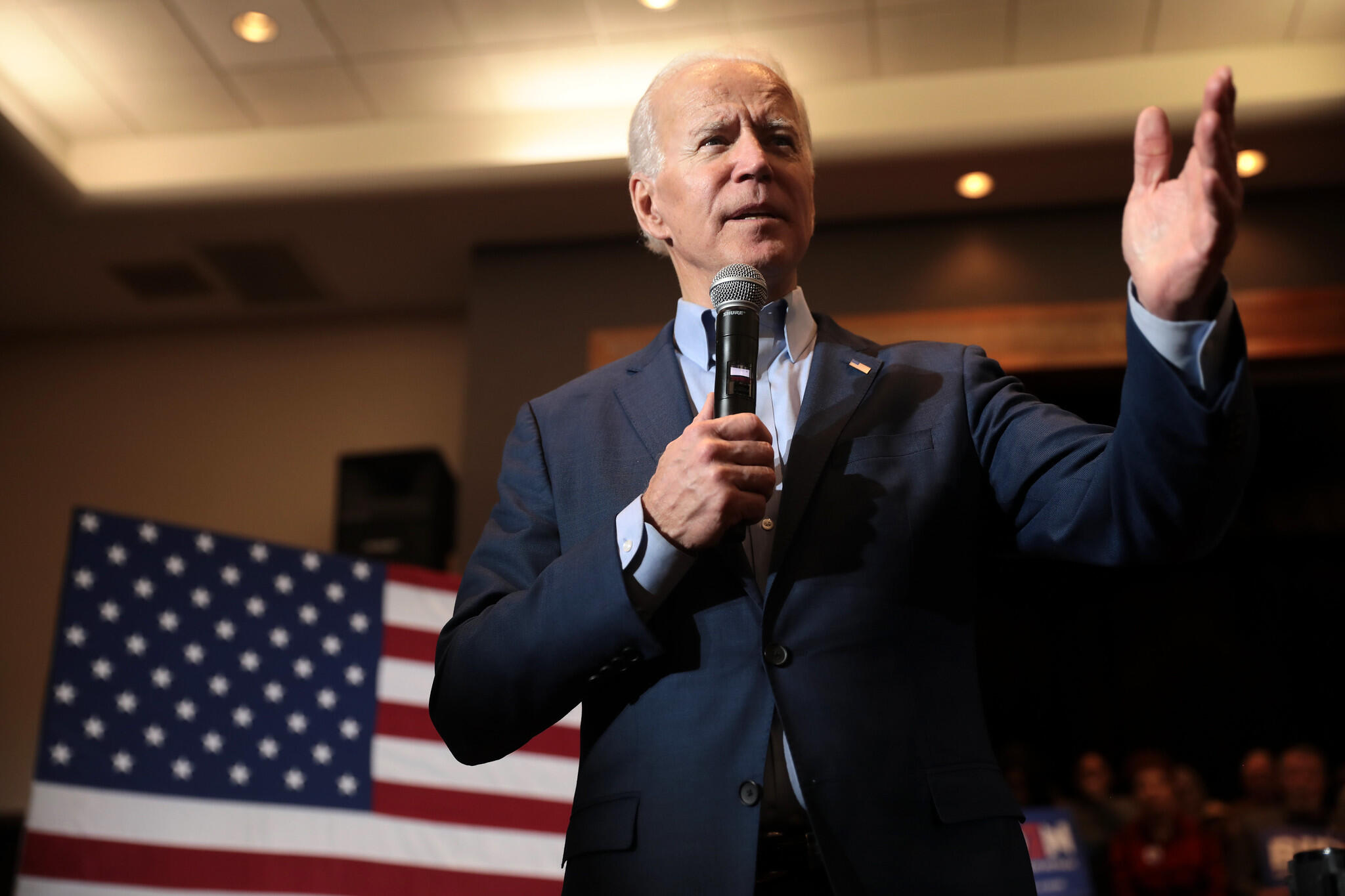 President Joe Biden speaks into a microphone in his hand and gestures with his other hand while standing in front of an American flag