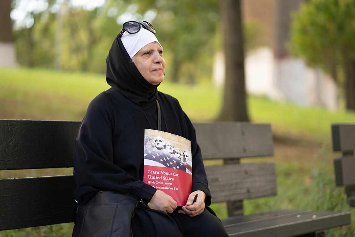 Maha al-Obaidi sits on a park bench looking off into the distance and holding a U.S. citizenship test study book.