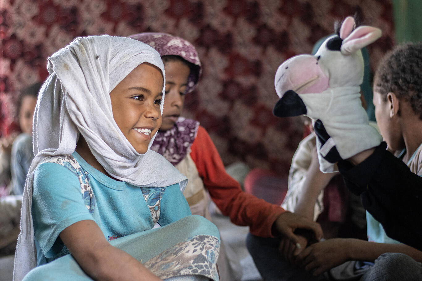 A little girl smiles and looks at a cow puppet while sitting on the ground with other children in Yemen