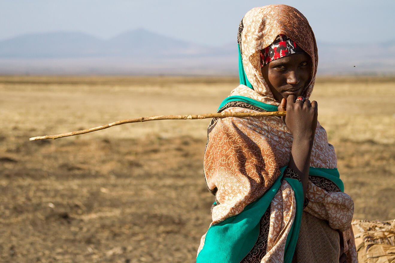 A teenage girl, holding a stick and looking at the camera, stands in a drought affected very dry landscape with mountains in the distance. 